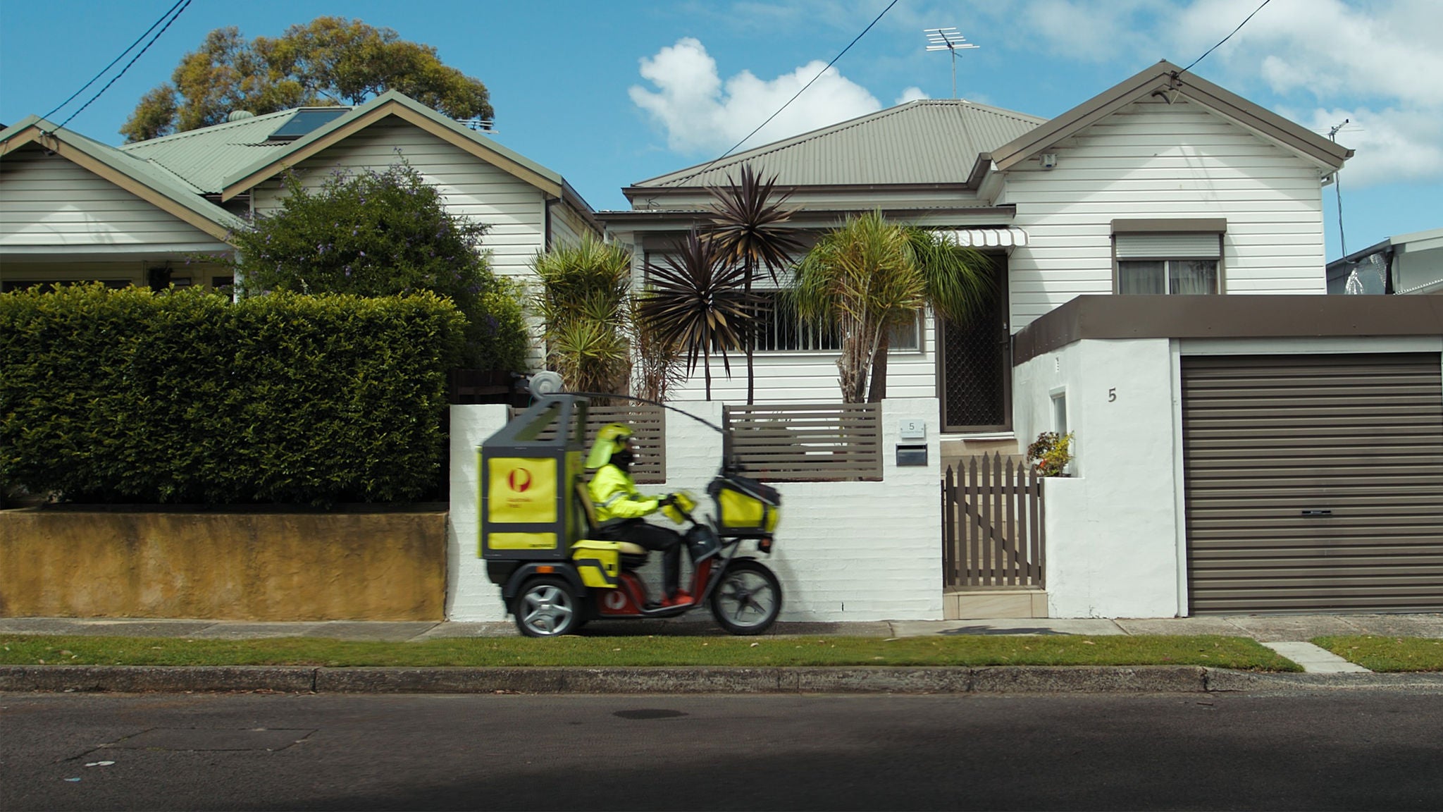 A delivery driver wearing a high-visibility yellow jacket rides a scooter past a white suburban house. The house features a modern facade, palm plants, and a closed garage door. The driver is in motion on the street, emphasizing urban delivery services. The sky is partly cloudy, and the scene is set in a quiet residential neighborhood.