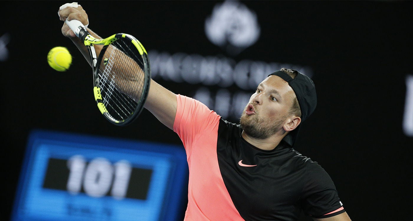 A male tennis player is captured mid-serve on an indoor court, wearing a black and pink athletic outfit. The action shot highlights the motion of the racket and ball, with a digital scoreboard in the background displaying the number 101. The setting suggests a competitive sports event with dramatic lighting and a focused atmosphere.