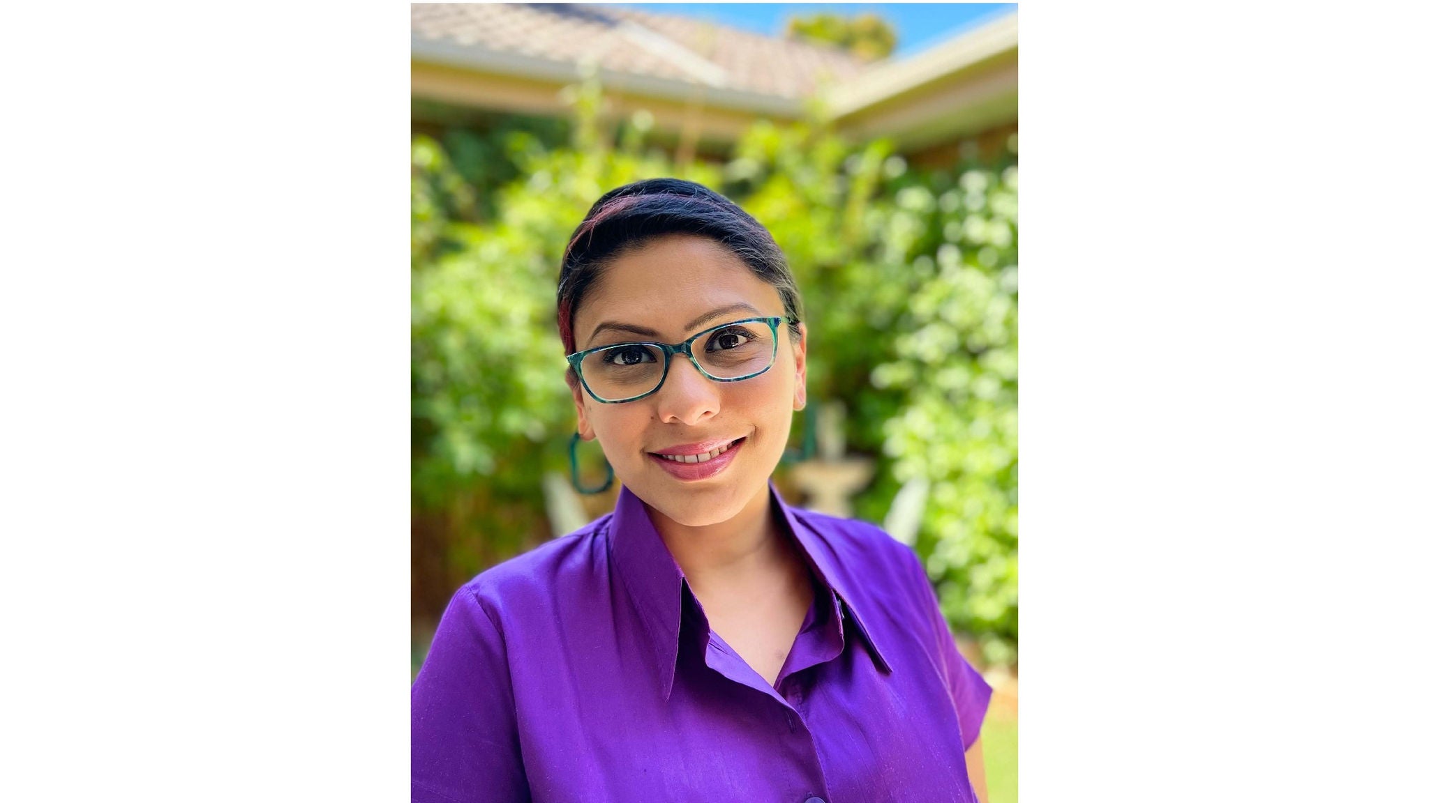 A person wearing a vibrant purple collared shirt is pictured standing outside in a lush garden setting. The background features greenery and part of a house with a tiled roof, suggesting a residential environment. The image is brightly lit, indicating a sunny day, and the overall mood is relaxed and casual.