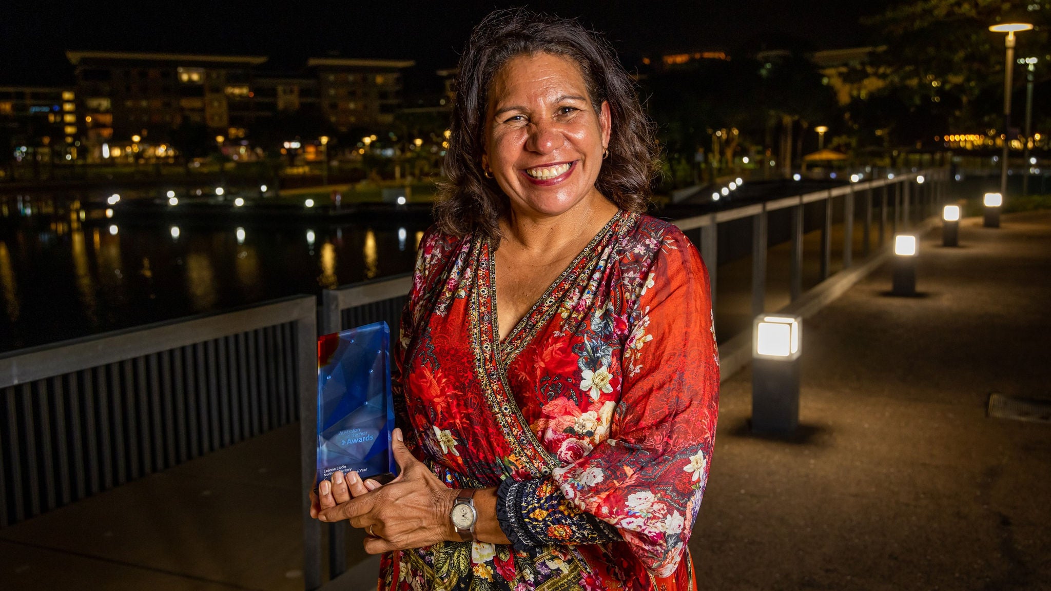 A woman stands outdoors at night, holding a blue glass award. She is dressed in a vibrant red floral dress with intricate patterns. The setting features a well-lit walkway beside a body of water, with modern buildings illuminated in the background. The atmosphere is celebratory and elegant.