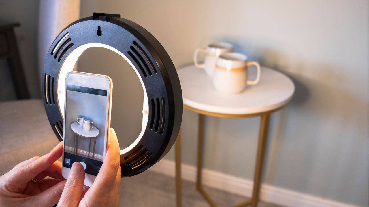 A person uses a smartphone and a circular ring light to photograph two white mugs on a small round table. The setting appears to be indoors with soft, neutral-colored walls and gold-accented furniture. The scene highlights a casual, creative activity with a focus on content creation or product photography. The lighting is bright and even, enhancing the clarity of the objects. No visible text or numbers are present in the image.