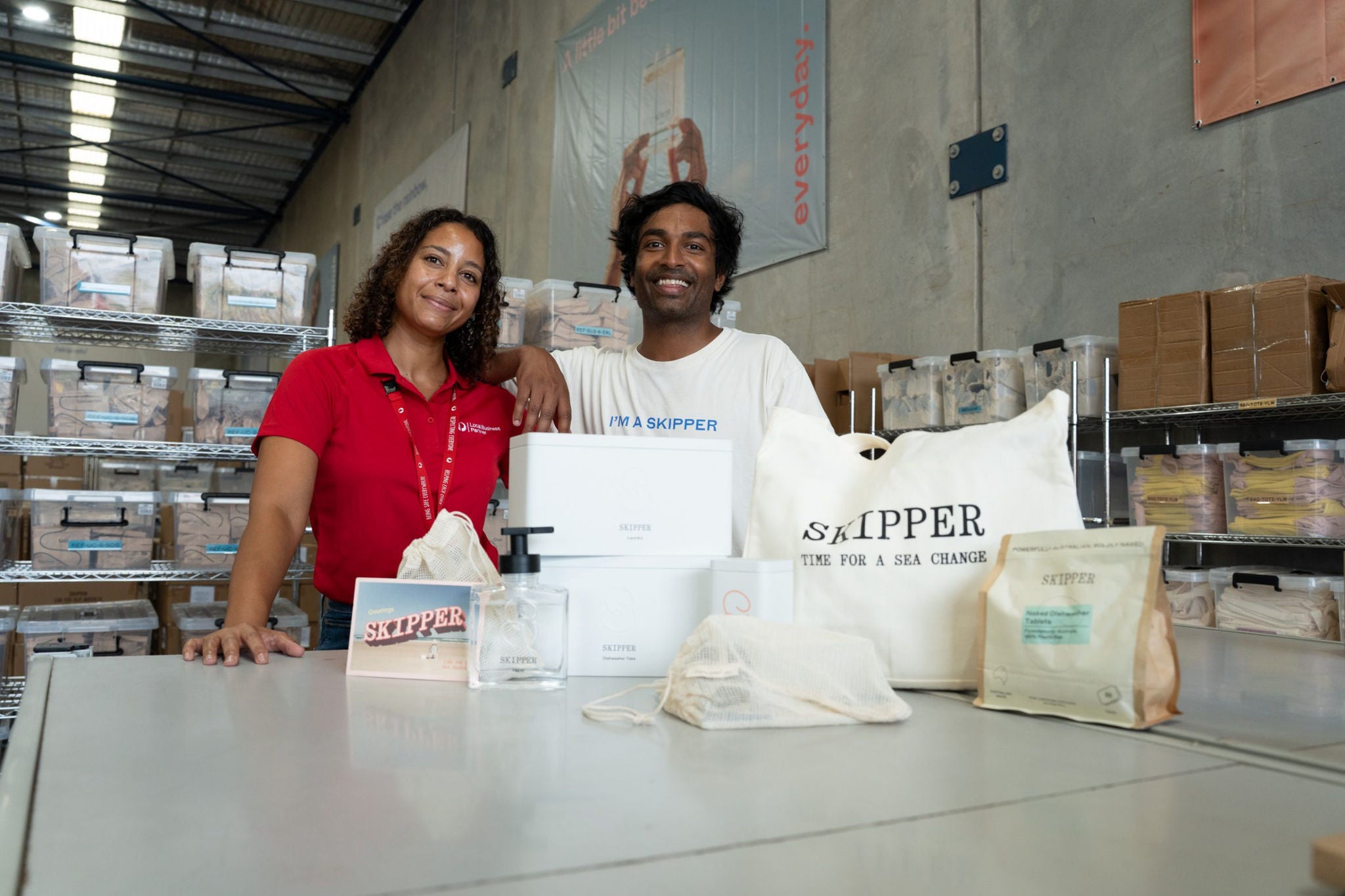 Two people stand behind a table in a warehouse filled with shelves and cardboard boxes. On the table, various Skipper items are arranged, including a tote bag, boxes, a pump bottle, pouches, and a small card. The workers wear casual clothing and lanyards, suggesting a professional or fulfillment setting. The industrial interior and organized inventory emphasize an e-commerce or small business environment.