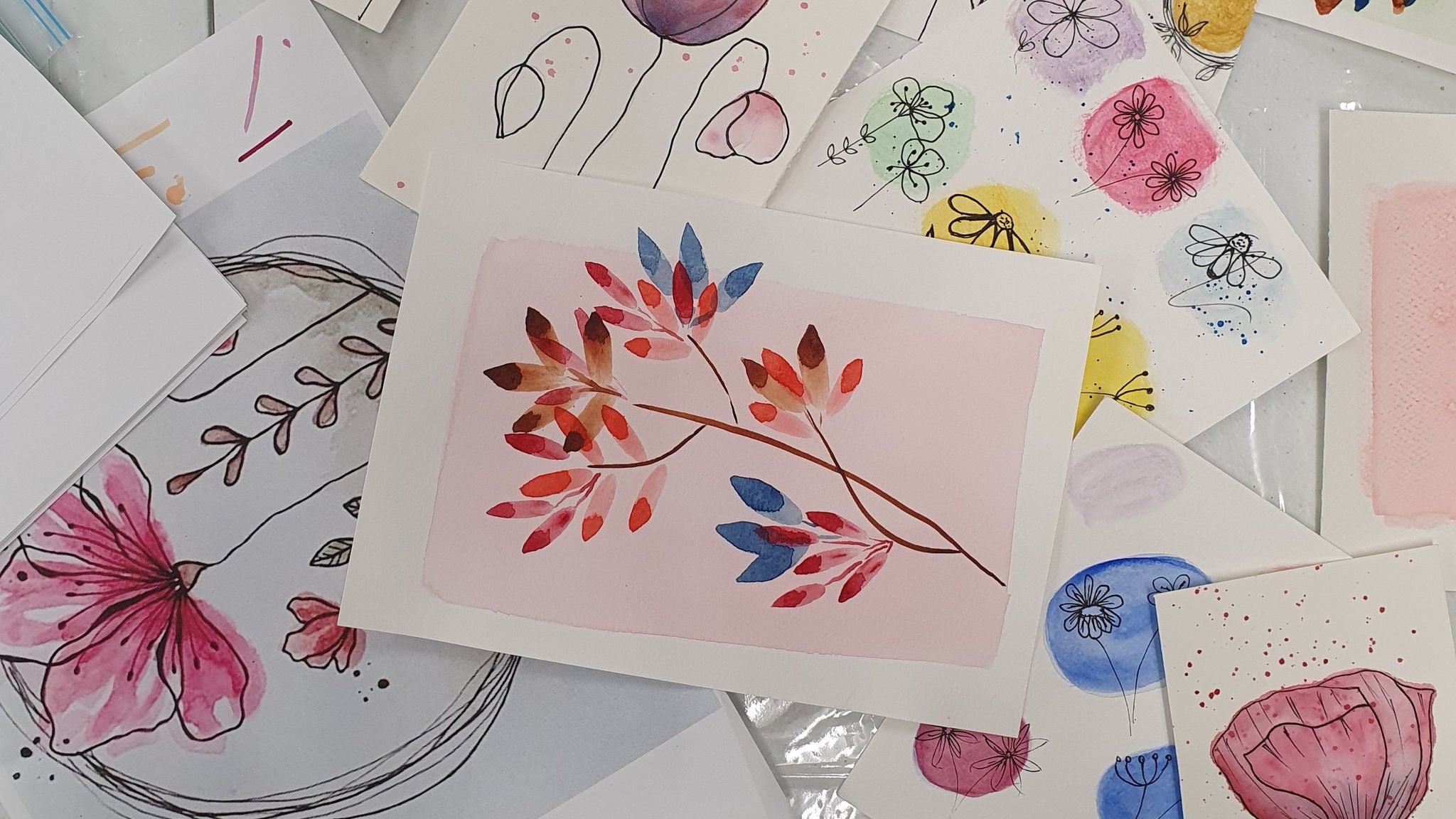 A collection of watercolor and ink drawings is spread out on a flat surface. The main subject is a colorful botanical branch painting on pink paper, surrounded by various floral sketches and abstract flower designs. The scene features soft pastel tones and hand-drawn artistic styles, with no visible text or numbers.