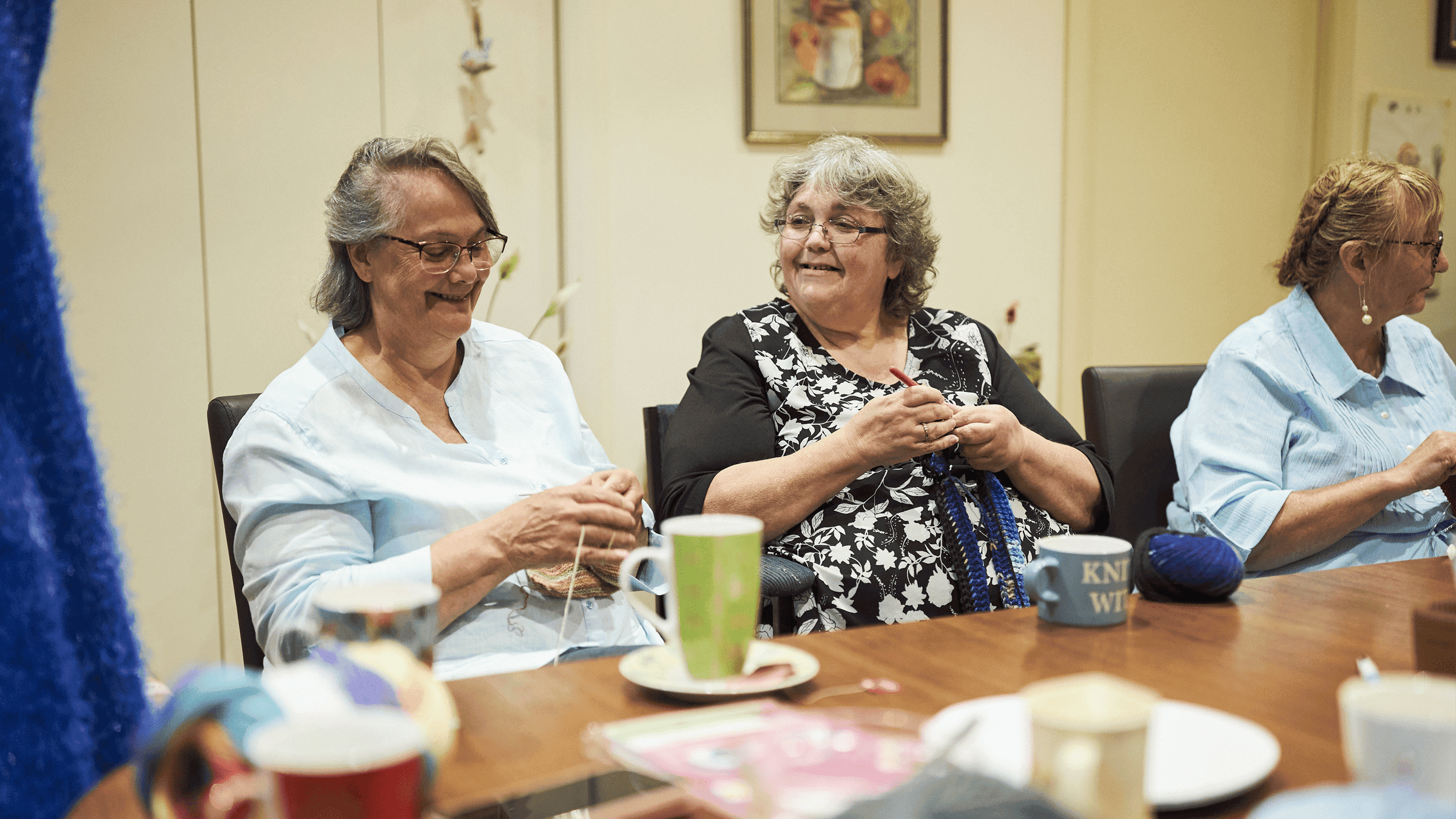 Three older women are seated around a wooden table, engaged in knitting activities. The setting appears to be a cozy indoor community or social room, with mugs, yarn, and knitting supplies visible on the table. The atmosphere is relaxed and social, with warm lighting and casual attire. A mug with the word 'KNIT' is clearly visible among the items. The scene conveys a sense of community and shared hobby.