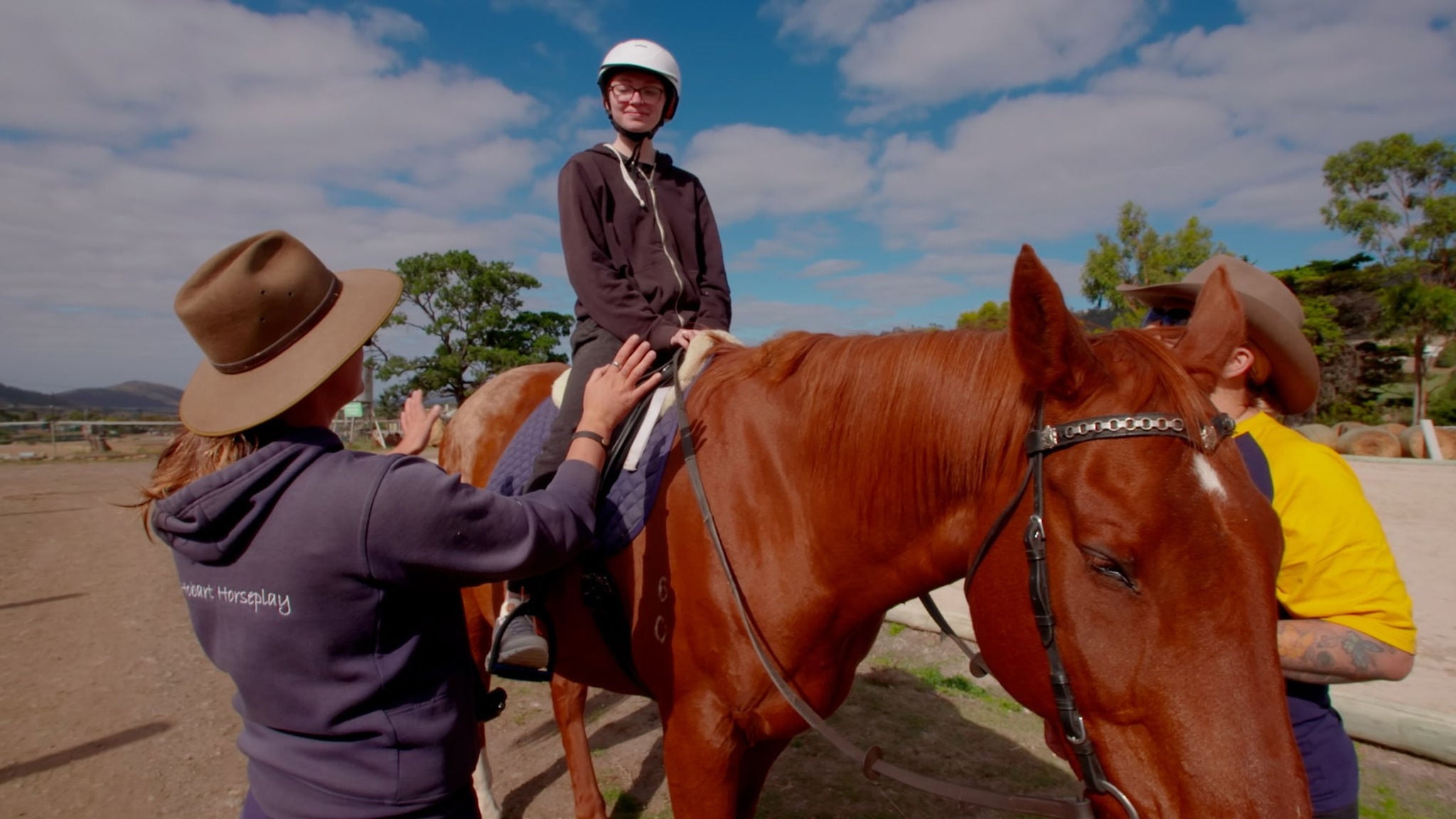 A young person sits on a brown horse while two adults provide guidance and support. The scene takes place outdoors under a partly cloudy sky, with trees and fencing in the background. The instructors wear wide-brimmed hats and casual clothing, creating a relaxed and instructional atmosphere. The focus is on equestrian activity and teamwork in a rural setting.