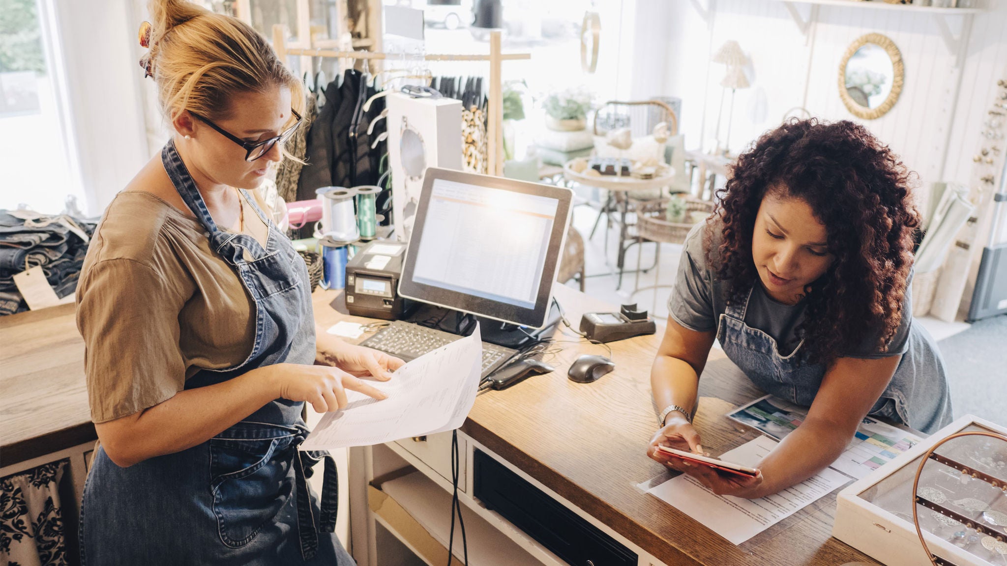 Two women are collaborating at a retail store counter, one reviewing paperwork while the other uses a smartphone. The setting features a computer monitor, cash register, and various retail items in a bright, modern shop environment. Both women are wearing aprons, suggesting they are employees. The workspace is organized, with natural light streaming in through large windows. No visible numeric values or identifiable brands are present.