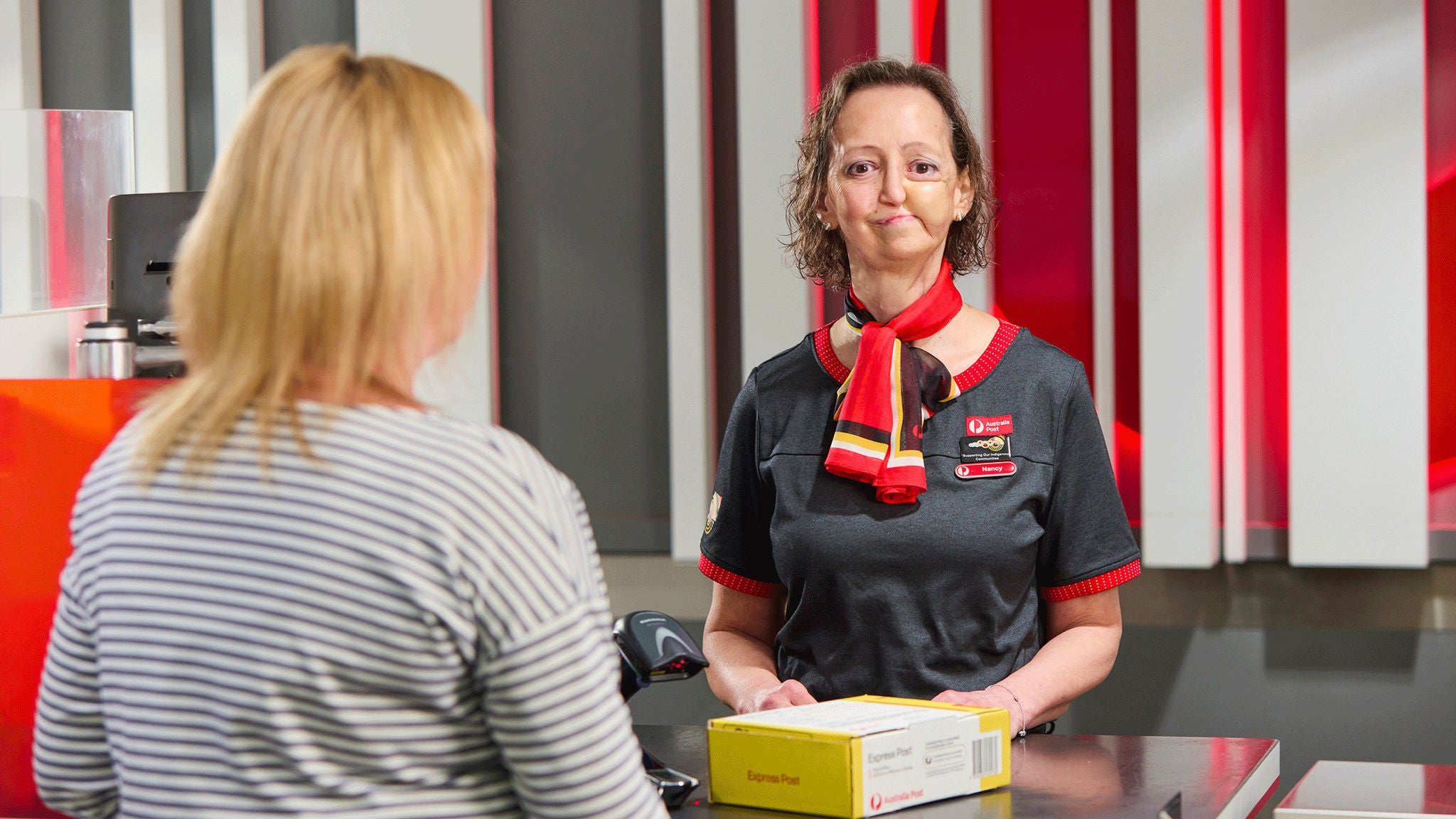 A postal worker in a uniform assists a customer at a modern service counter. The worker is holding a yellow and white parcel box, engaging in a transaction. The setting features a contemporary interior with red, white, and gray vertical panels. The scene conveys a professional and efficient atmosphere, with clear focus on customer service and package handling.