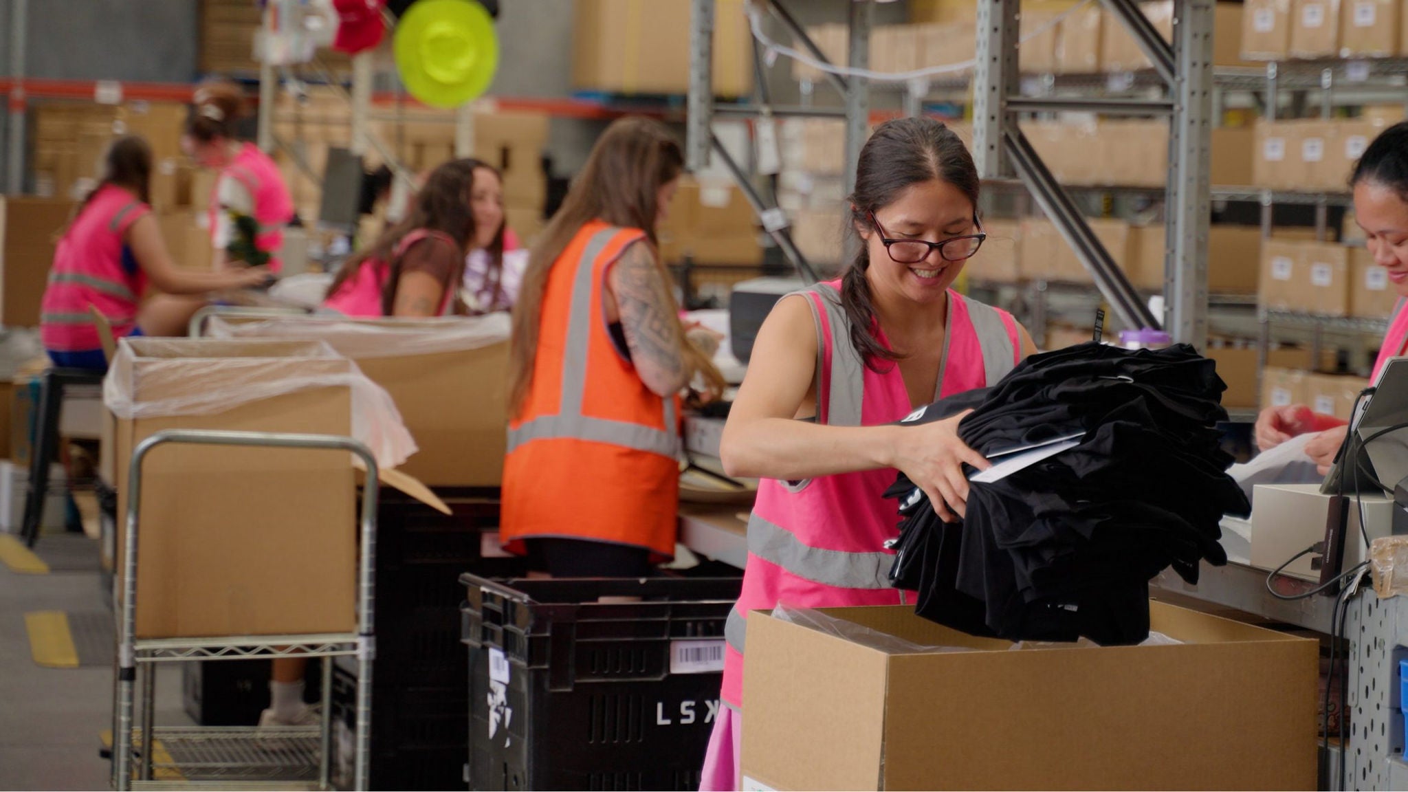 Several workers in high-visibility pink vests sort items in a busy warehouse. A woman in the foreground stacks folded black clothing near large cardboard boxes and plastic crates. Shelving units filled with brown cartons line the background, suggesting an organized distribution or fulfillment center. The scene conveys teamwork, logistics, and efficient order processing in an industrial environment.