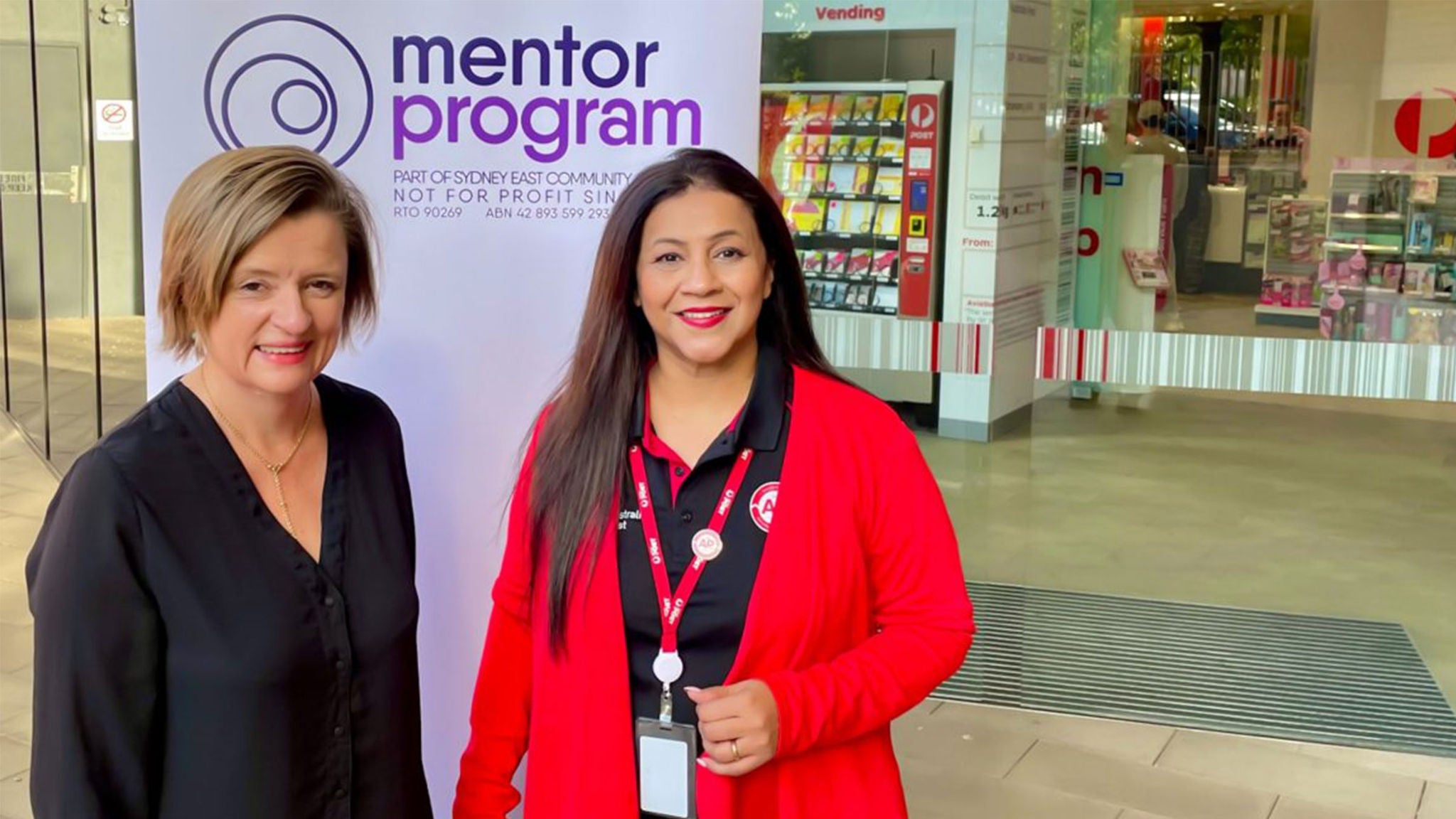 Two women stand in front of a sign reading 'mentor program' in a modern indoor setting. The background includes a vending machine, glass doors, and a display of brochures or magazines. Both women are wearing business casual attire, one in black and one in a red cardigan with a lanyard. The atmosphere suggests a professional or educational event.