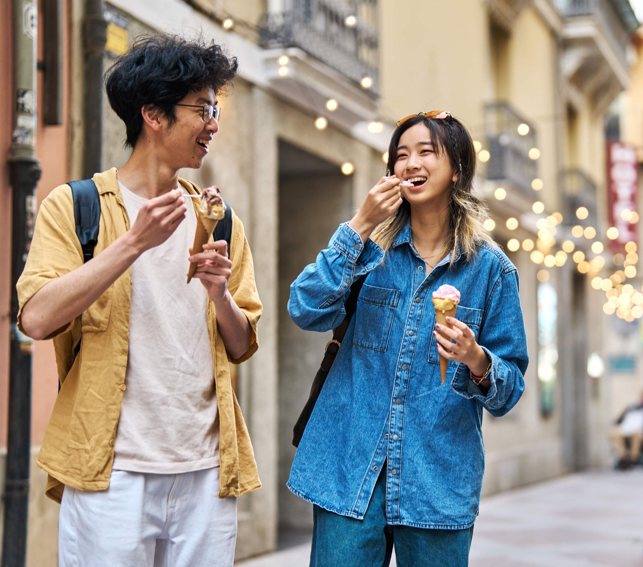 Two young adults are seen eating ice cream cones while walking along a charming city street. The setting features warm lighting and blurred string lights, suggesting a relaxed, urban atmosphere. Both individuals are casually dressed, one in a yellow shirt and the other in a blue denim jacket, each carrying a backpack. The scene conveys a casual, cheerful mood in an outdoor urban environment.