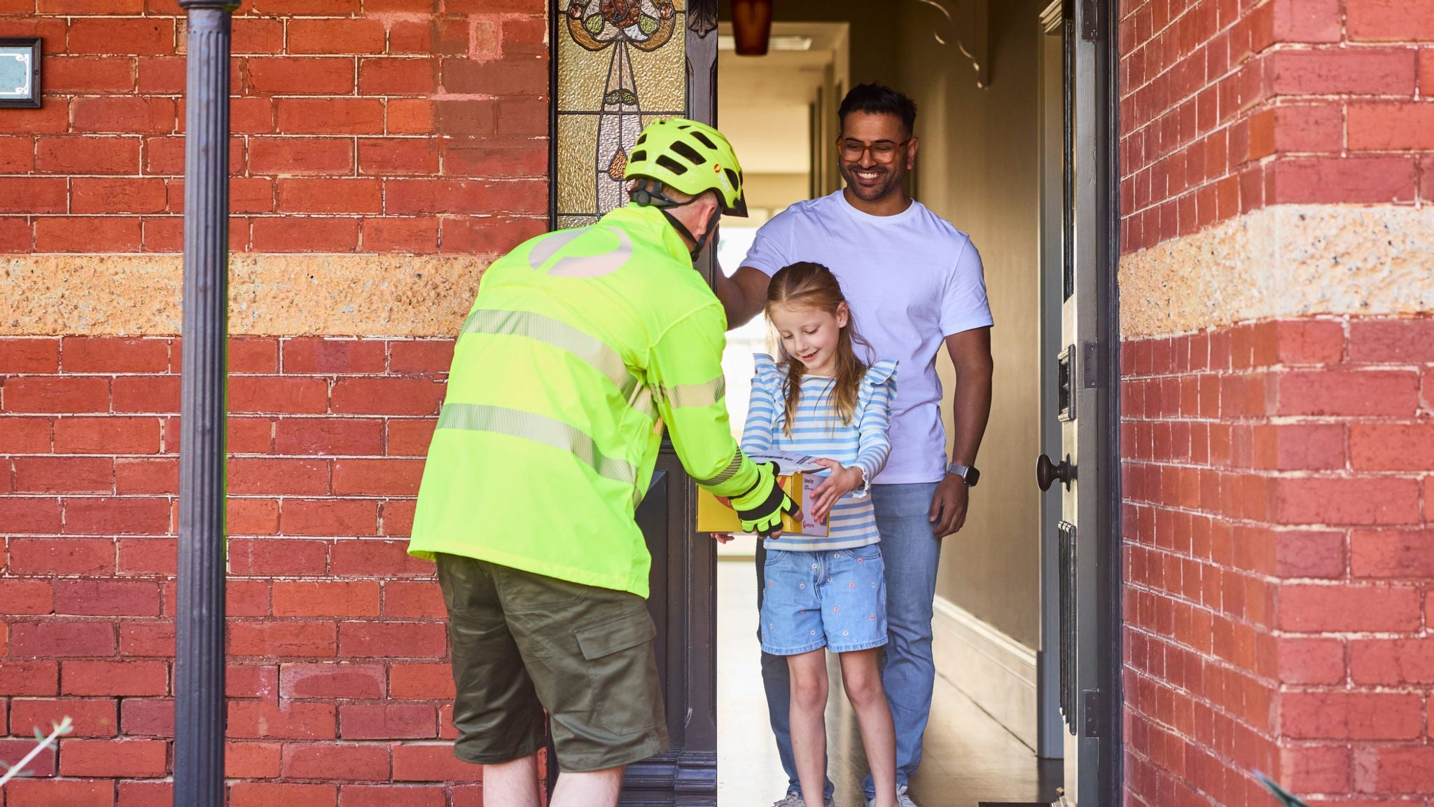 A delivery worker in a bright safety jacket and helmet hands a parcel to a young girl standing at a front door. The girl is accompanied by an adult man, likely a parent, in casual clothing. They stand in the doorway of a brick house with a decorative glass panel and warm interior lighting. The scene suggests a friendly home delivery interaction in a suburban neighborhood.