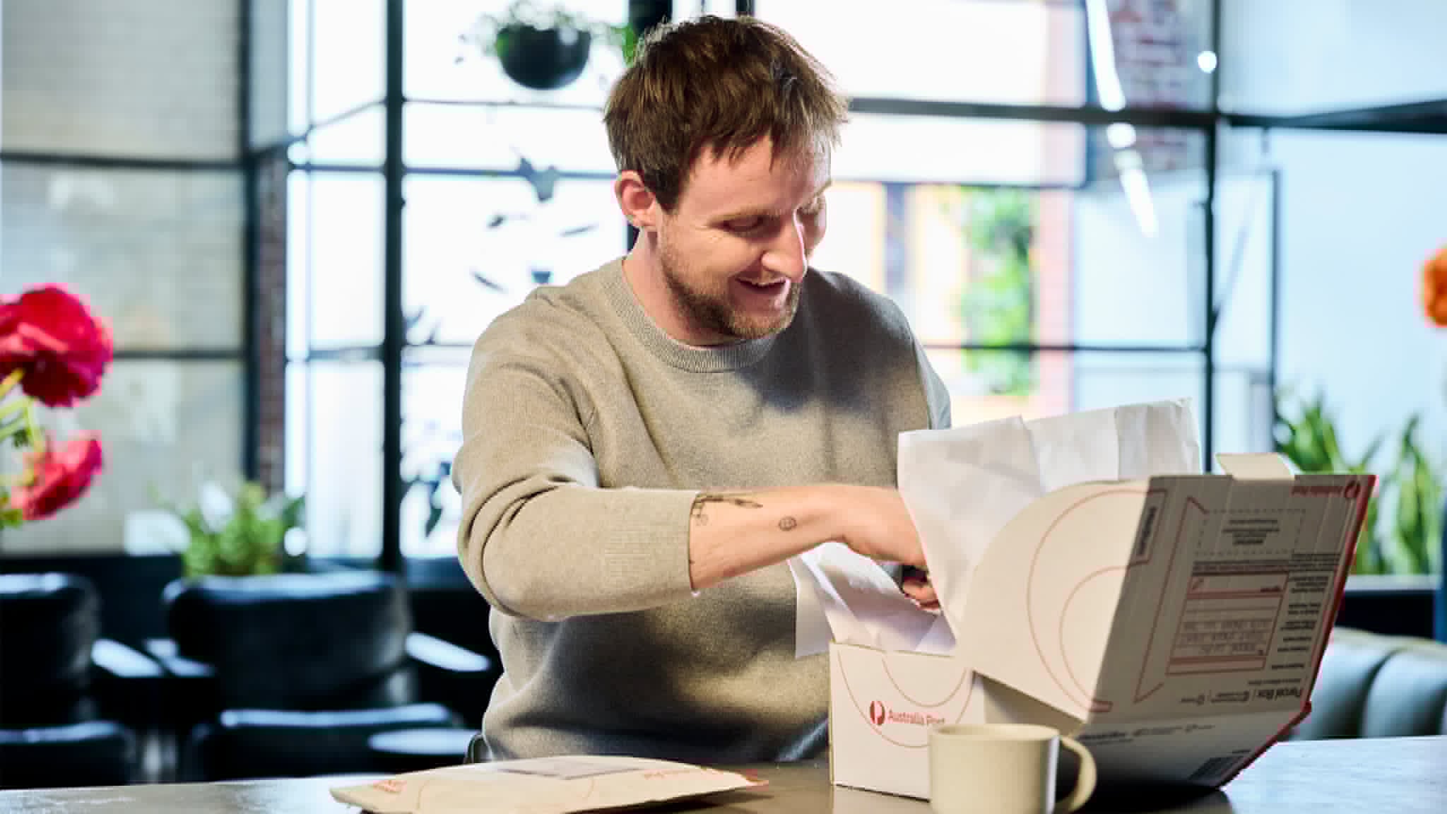 A young man sits at a table opening a white shipping box. The scene appears to be indoors in a bright, modern space with large windows and plants in the background. Tissue paper and mailing materials are visible as he looks into the package. A mug and an envelope or document lie on the table, suggesting a casual, everyday moment of receiving mail.