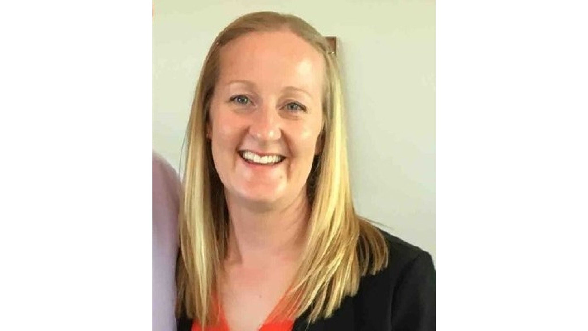 A woman with straight, shoulder-length blonde hair is pictured against a plain light background. She is wearing a black blazer over an orange top, suggesting a professional or business setting. The image is well-lit with neutral tones and a simple composition.