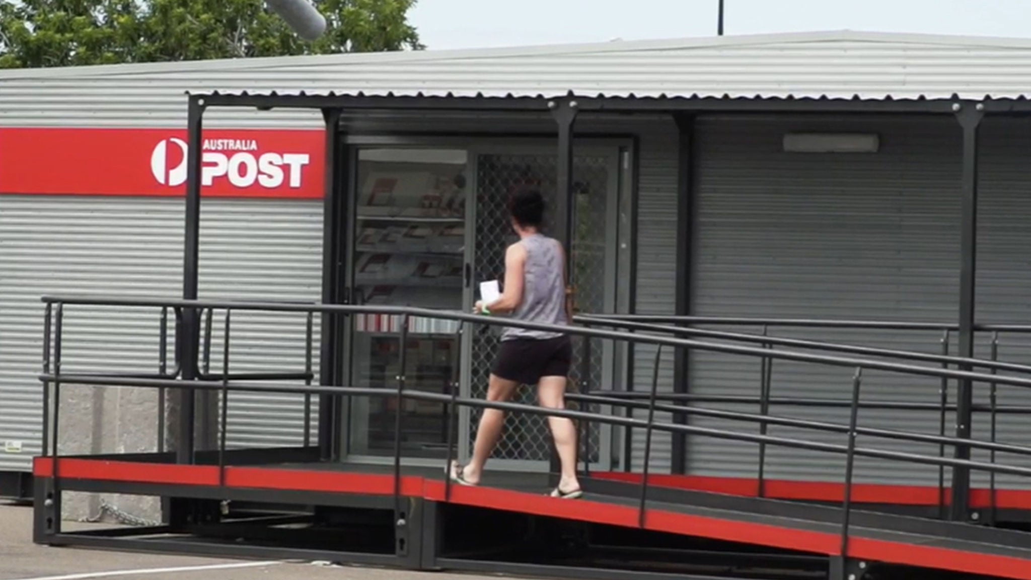 A person is walking up a ramp towards the entrance of an Australia Post office. The setting is outdoors, with a modern, corrugated metal building and clear signage. The individual appears to be an adult, casually dressed, and carrying a small package. The scene is well-lit, suggesting daytime, and the overall mood is practical and everyday. The Australia Post logo and name are clearly visible above the entrance.