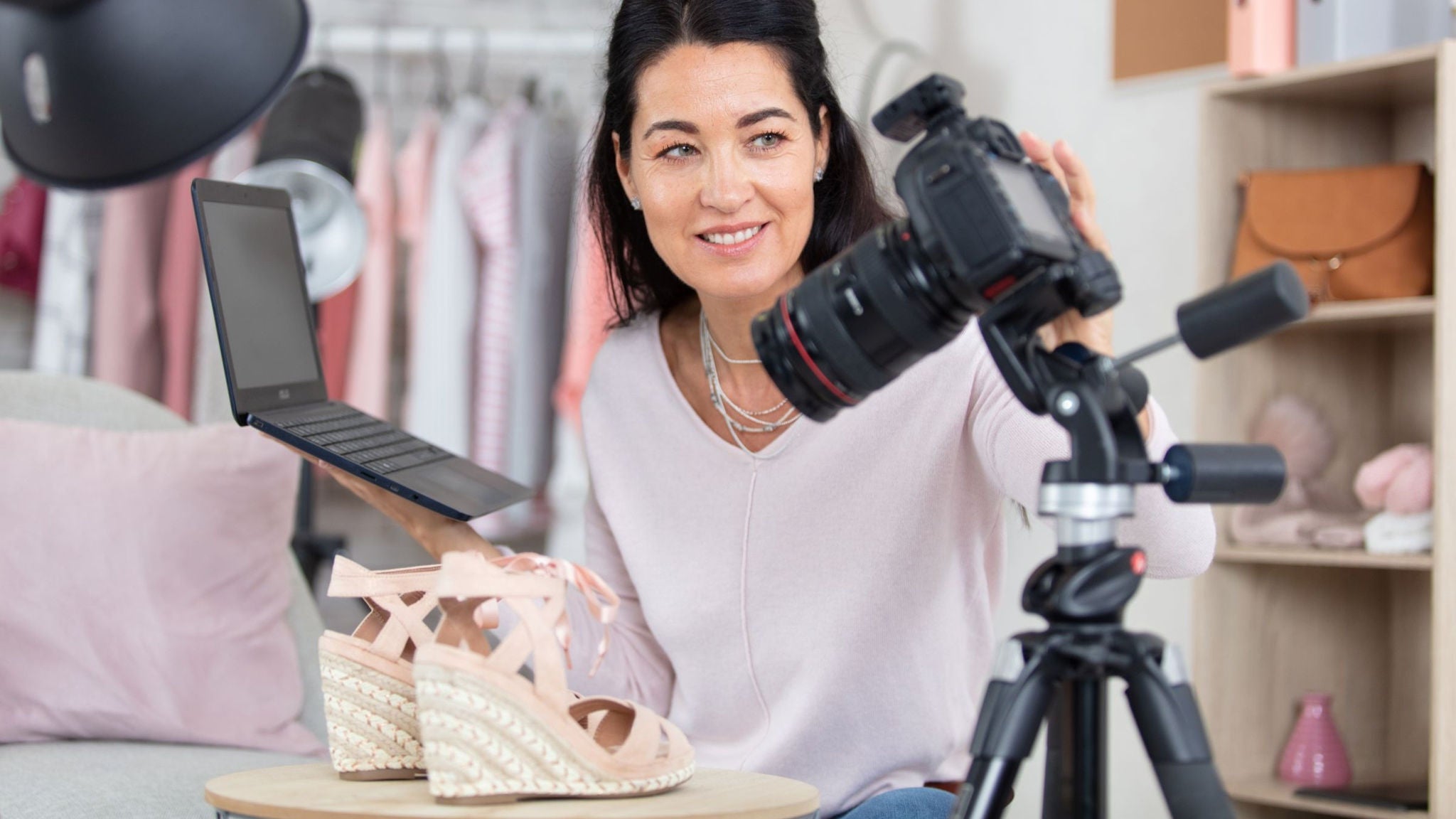 A young woman records a video review of pink wedge sandals using a digital camera on a tripod. She holds an open laptop while sitting in a stylish room with clothing racks and soft furnishings in the background. The scene suggests content creation or live streaming focused on fashion products. Soft pastel colors and casual decor create a relaxed, modern atmosphere.