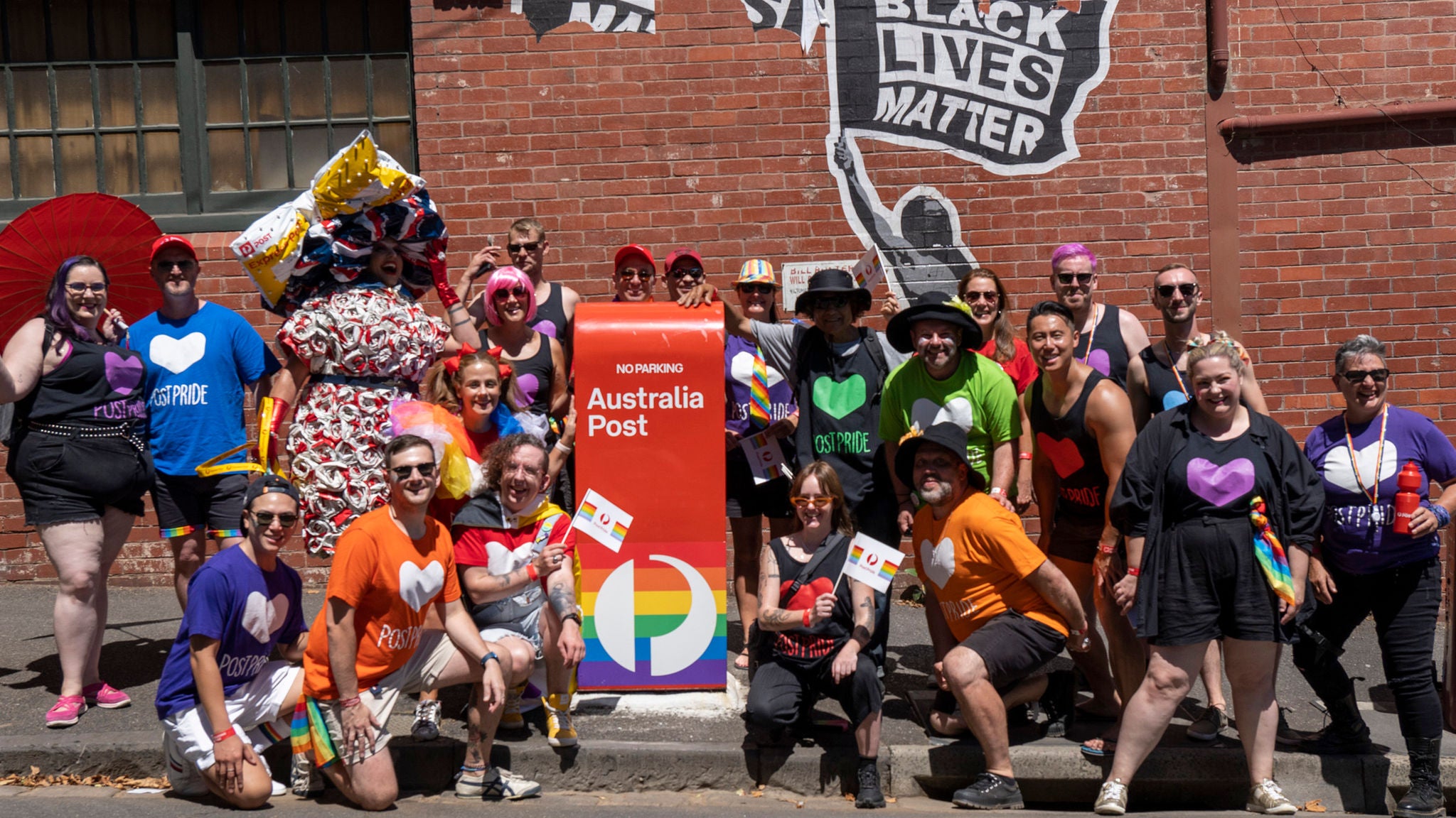 A diverse group of people gathers outdoors around a bright red Australia Post mailbox decorated with a rainbow circle. The group is dressed in colorful clothing, many wearing shirts with heart designs and holding small rainbow flags. The setting features a brick wall with bold street art, including the phrase 'BLACK LIVES MATTER.' The mood is festive and inclusive, with a strong emphasis on LGBTQ+ pride and community.