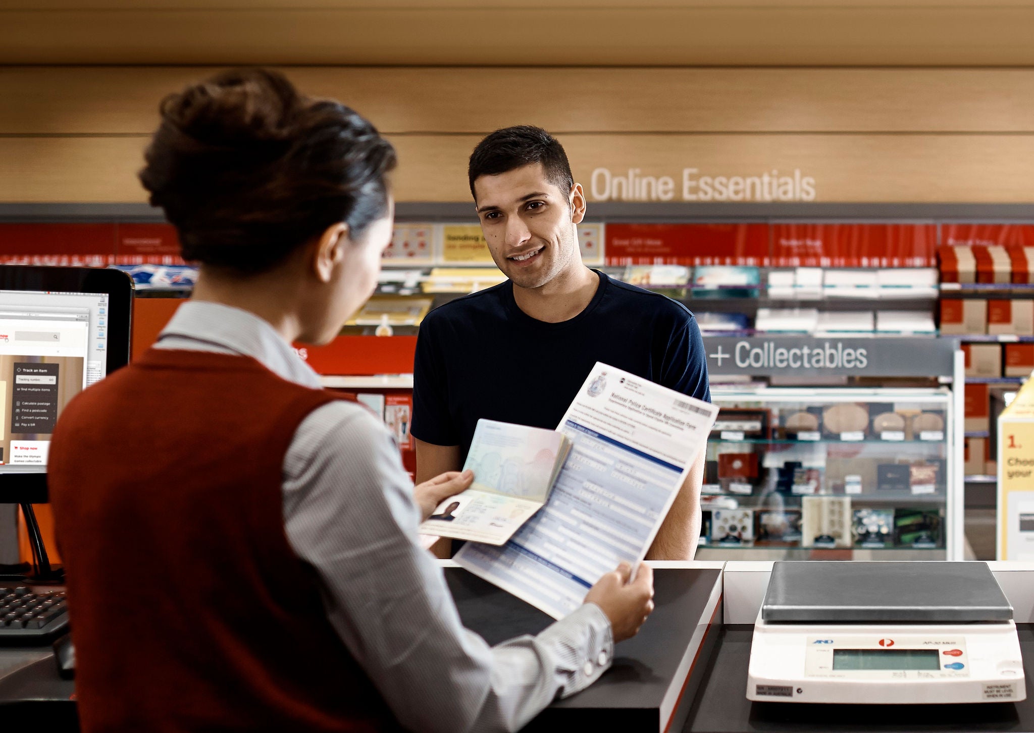 A customer stands at a service counter handing over documents and an identification card to a retail clerk. The interaction takes place in a brightly lit store with shelves of products and signage in the background. A digital scale and computer monitor are visible on the counter, suggesting a shipping or postal transaction. The scene conveys an organized, professional environment focused on customer service.