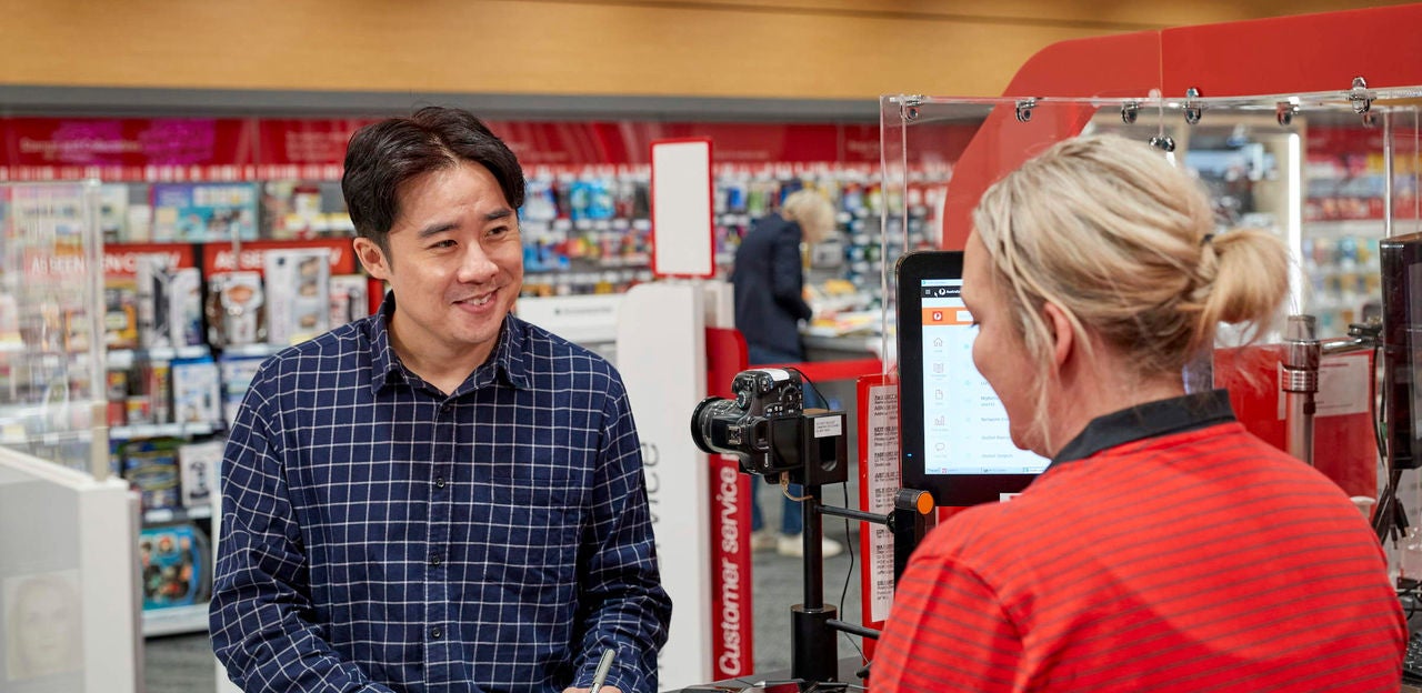 A man in a blue checkered shirt stands at a customer service counter in a retail store, engaging with a staff member in a red uniform. The setting features shelves stocked with various products and a digital screen at the counter. The environment is brightly lit, with a modern and organized appearance. A camera device is positioned on the counter, possibly for identification or transaction purposes.