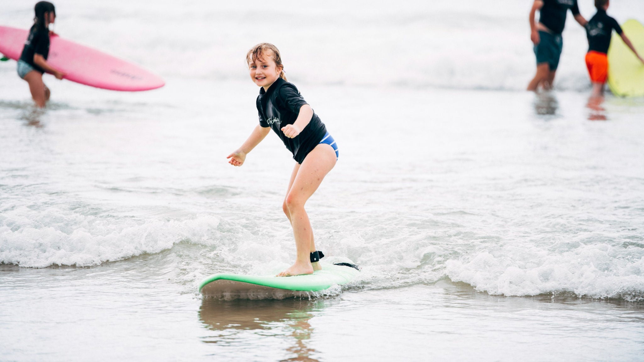 A young person is learning to surf, balancing on a light green surfboard in shallow ocean waves. The setting is a lively beach with other people and surfboards visible in the background. The mood is energetic and playful, with overcast lighting and soft, muted colors. The scene captures a moment of focus and excitement during a surf lesson.