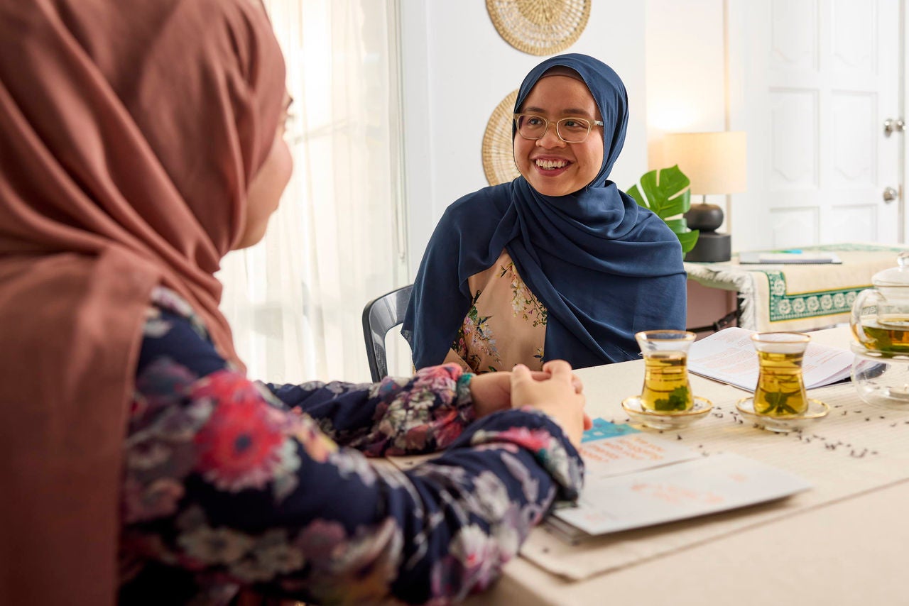 Two women wearing floral and solid-colored headscarves sit across from each other at a table in a bright, cozy room. The table is set with tea in clear glasses, documents, and a pen, suggesting a casual meeting or friendly conversation. The background features woven wall decor, a leafy plant, and soft natural lighting, creating a warm and inviting atmosphere.