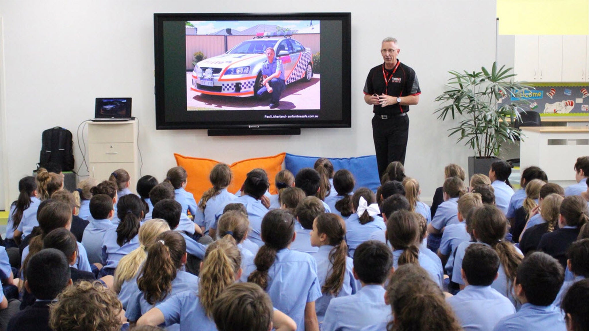 A police officer stands in front of a large screen, giving a presentation to a group of young school children seated on the floor. The screen displays an image of a police car, and the classroom setting includes colorful cushions and a potted plant. The children are wearing matching uniforms, and the atmosphere is educational and attentive.