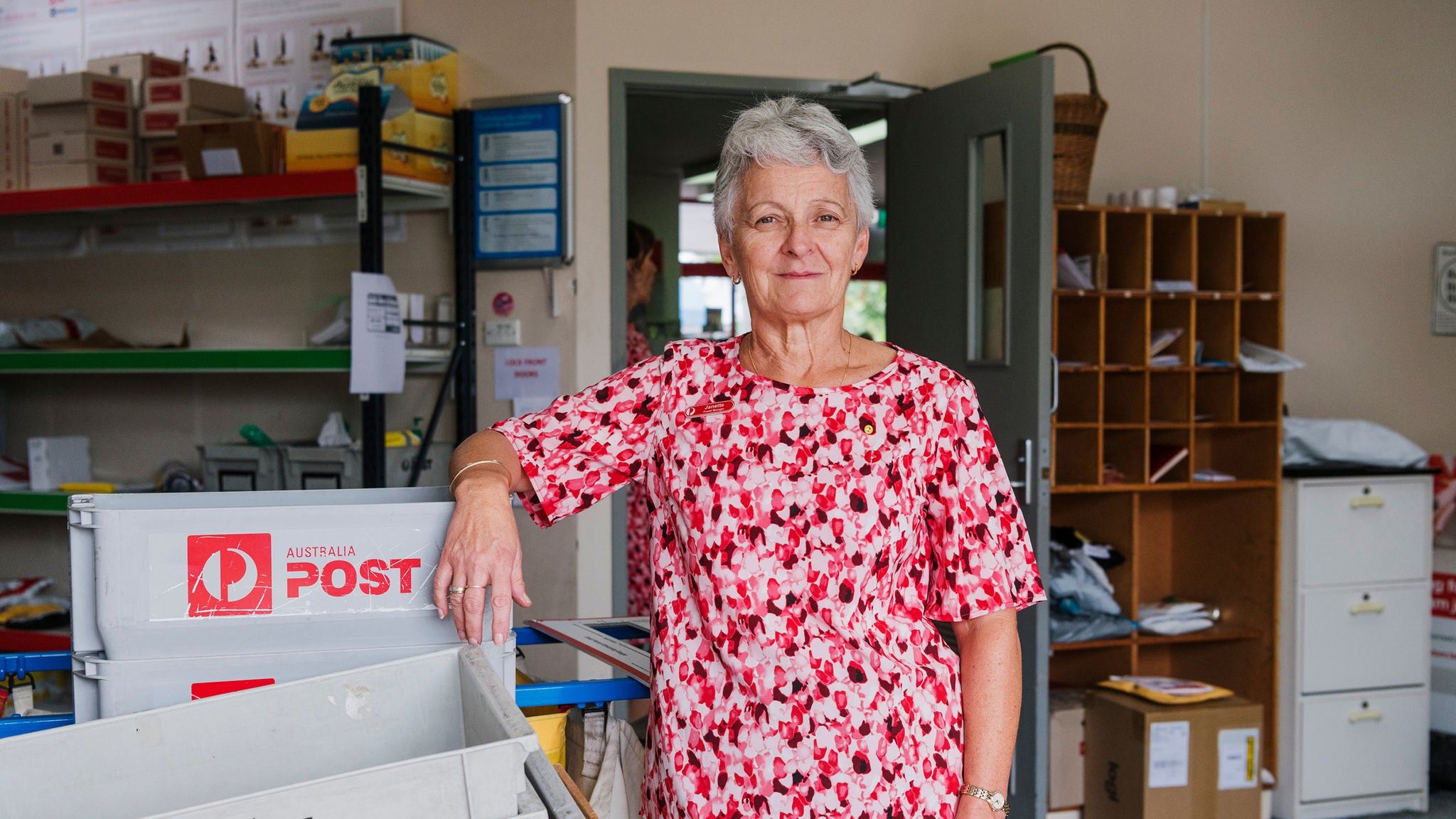 An older woman wearing a red and white floral shirt stands inside a post office, leaning on a stack of Australia Post mail crates. The setting features organized shelves, mail slots, and office supplies, indicating a busy postal environment. The Australia Post logo is clearly visible on the crates, and the workspace appears functional and well-used.