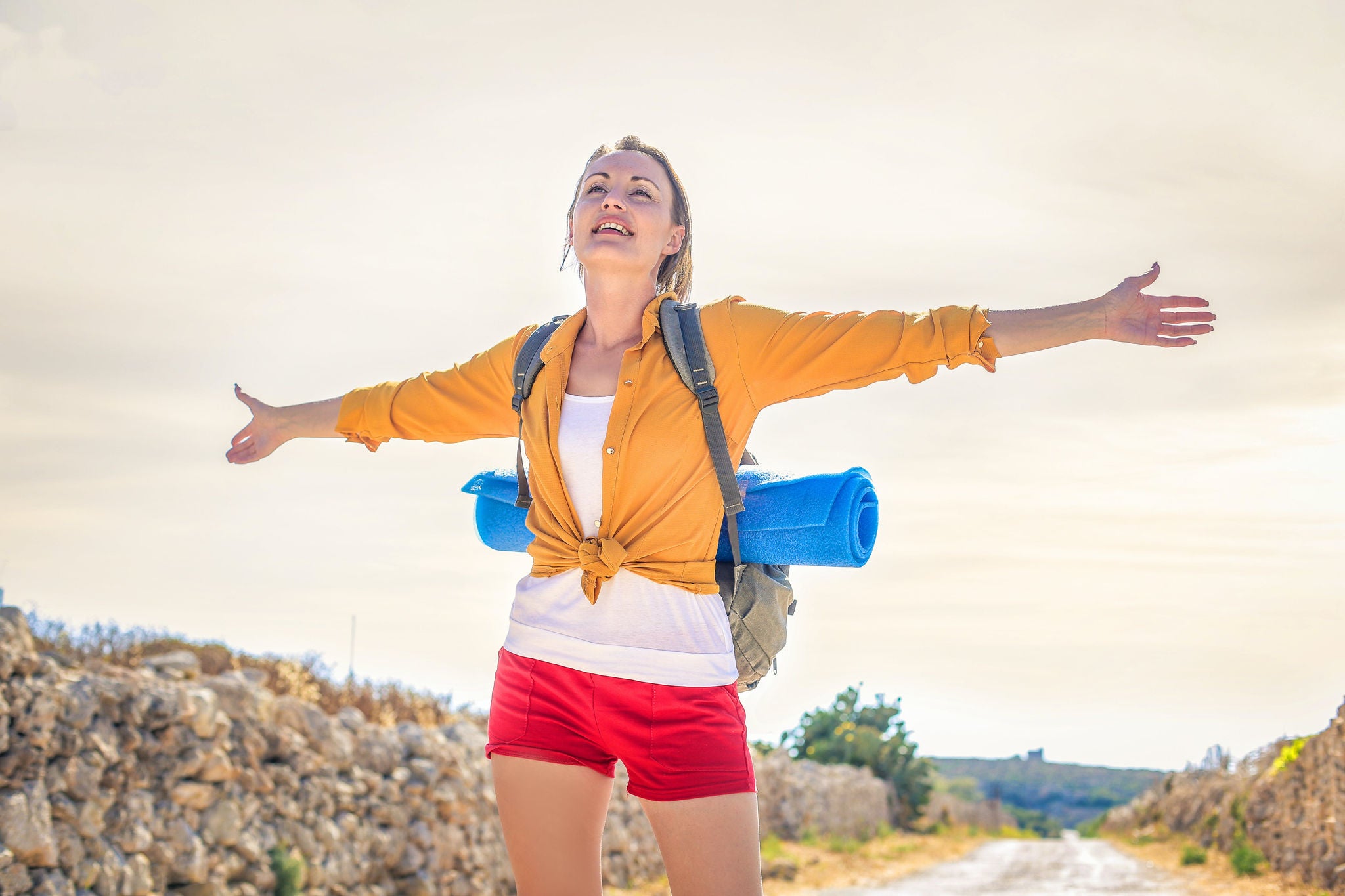 A woman stands on a rural path with arms outstretched, wearing a yellow shirt and red shorts. She carries a backpack and a blue rolled-up sleeping mat, suggesting she is on a hiking or camping trip. The background features a stone wall and open countryside under a bright sky. The image conveys a sense of freedom and adventure.