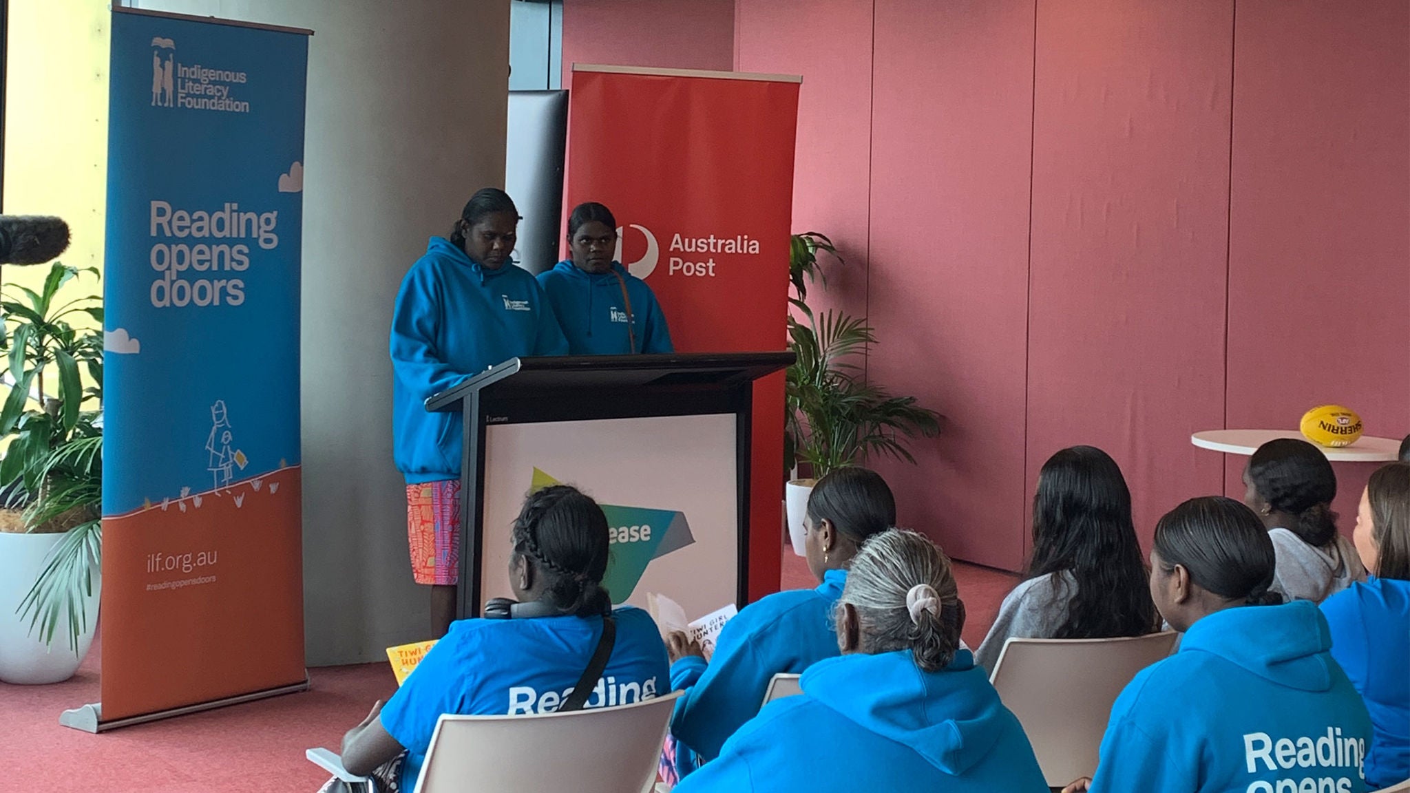 A group of young people, mostly girls, are gathered indoors for a literacy-focused event. Two youths stand at a podium, presenting to an audience wearing matching blue hoodies with 'Reading opens doors' printed on the back. Visible banners include 'Australia Post' and 'Reading opens doors,' emphasizing the event's educational theme. The setting is modern, with red and yellow accents and a casual, engaged atmosphere.
