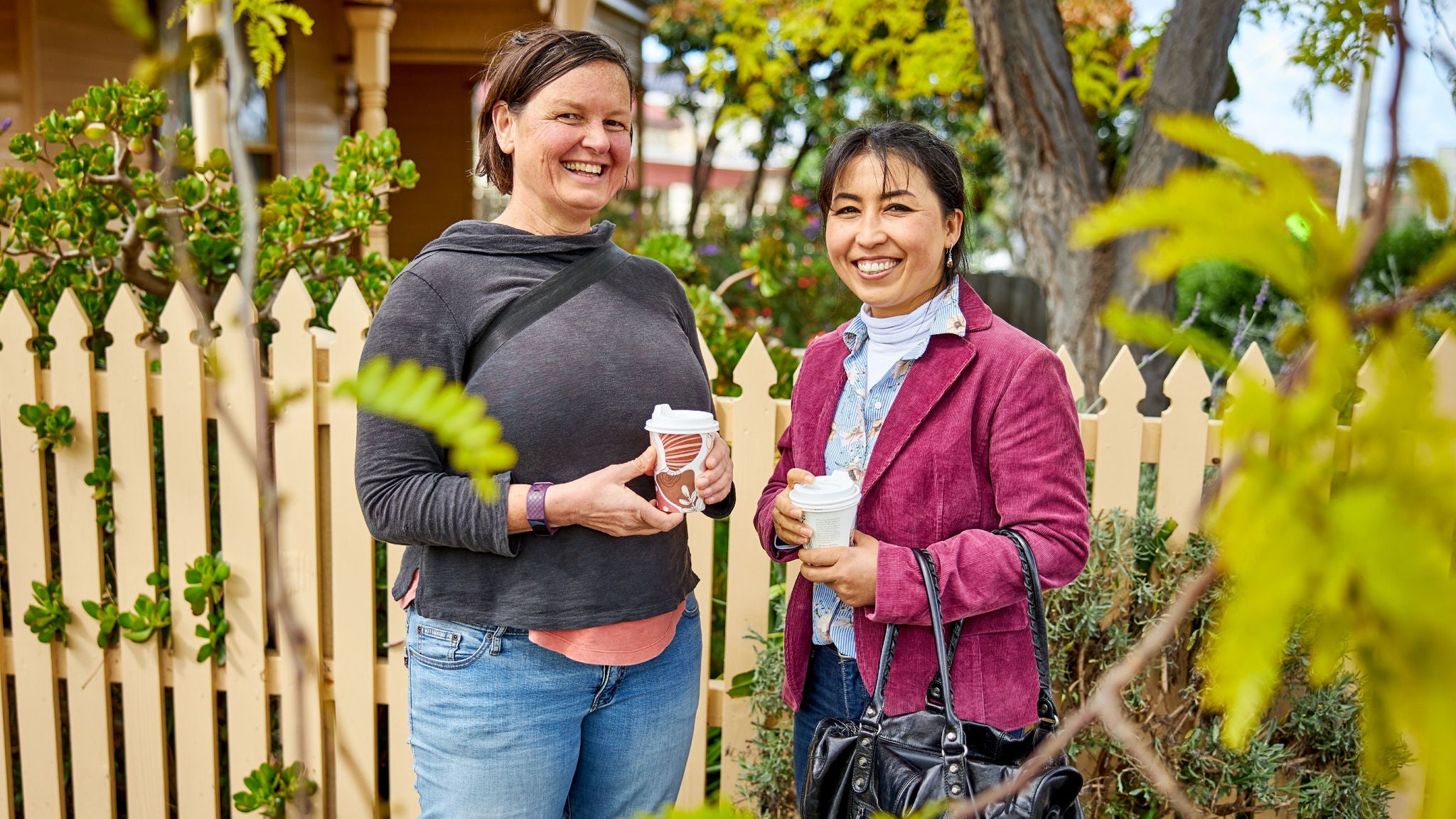 Two adults are standing outside in a residential area, each holding a takeaway coffee cup. They are positioned in front of a light-colored picket fence with greenery and trees in the background. One woman is wearing a dark top and jeans, while the other is dressed in a red jacket and holding a black handbag. The setting suggests a casual, relaxed atmosphere in a suburban neighborhood.