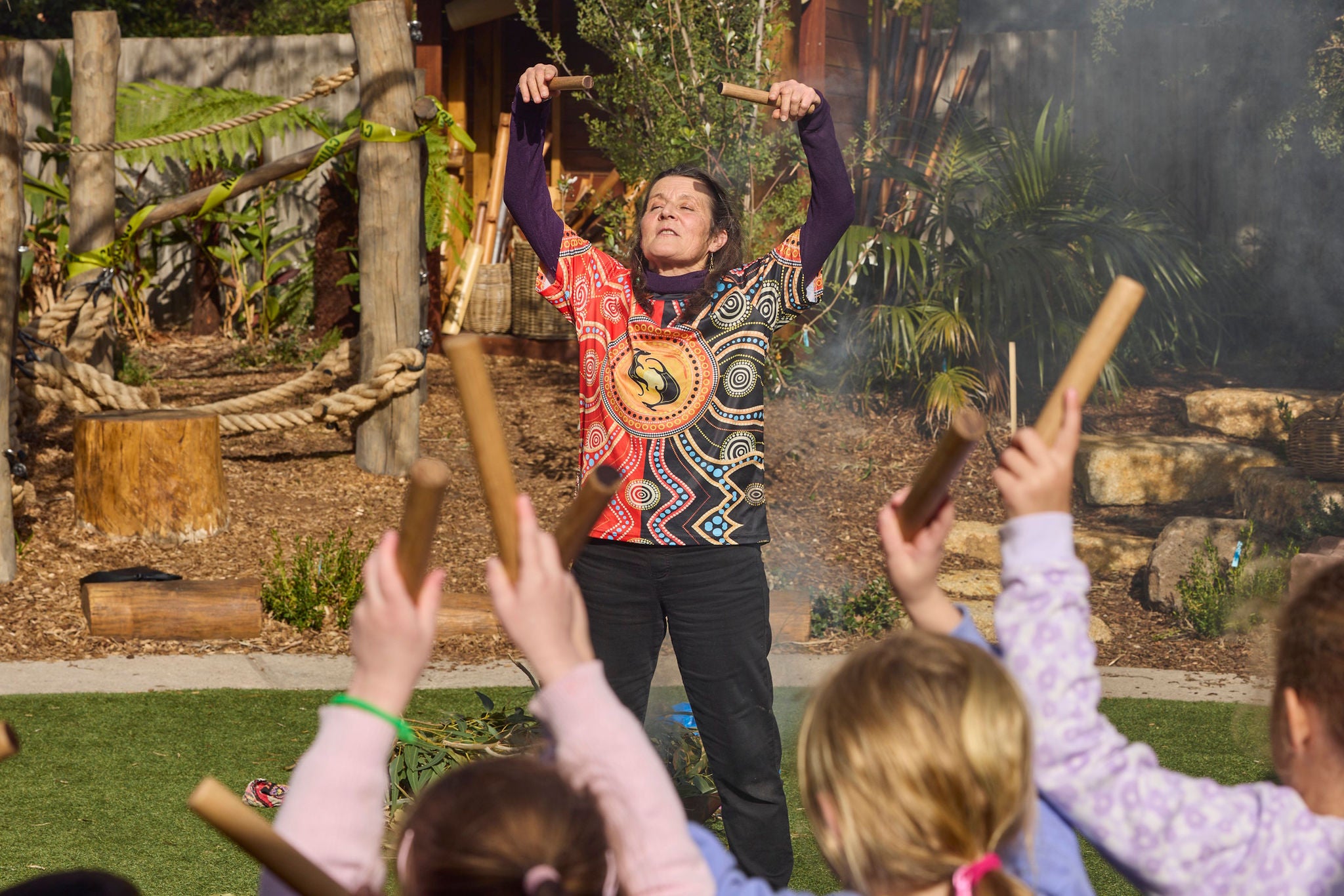 An adult instructor leads a group of young children in an outdoor music activity, holding wooden rhythm sticks above their head. The children, seated or standing in front, also hold up rhythm sticks, participating enthusiastically. The setting features natural elements like logs, plants, and ropes, suggesting a nature-inspired or educational environment. The instructor wears a colorful, patterned shirt, and the mood is lively and engaging.
