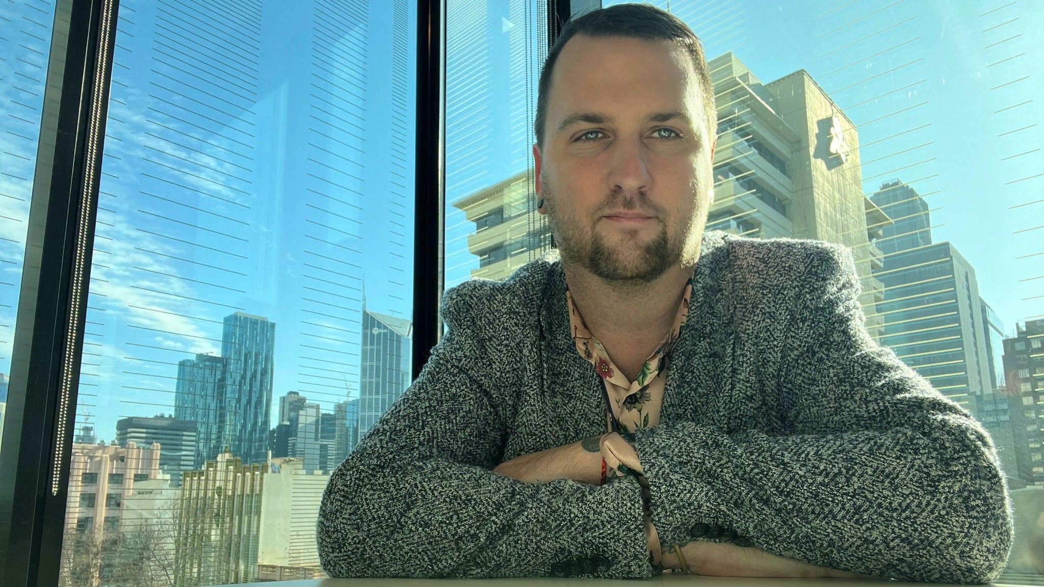A person sits at a table in a modern office space with large glass windows. The background features a clear view of a city skyline with tall buildings and blue sky. The individual is wearing a textured gray jacket and a patterned shirt. Sunlight streams through the windows, creating a bright and airy atmosphere.