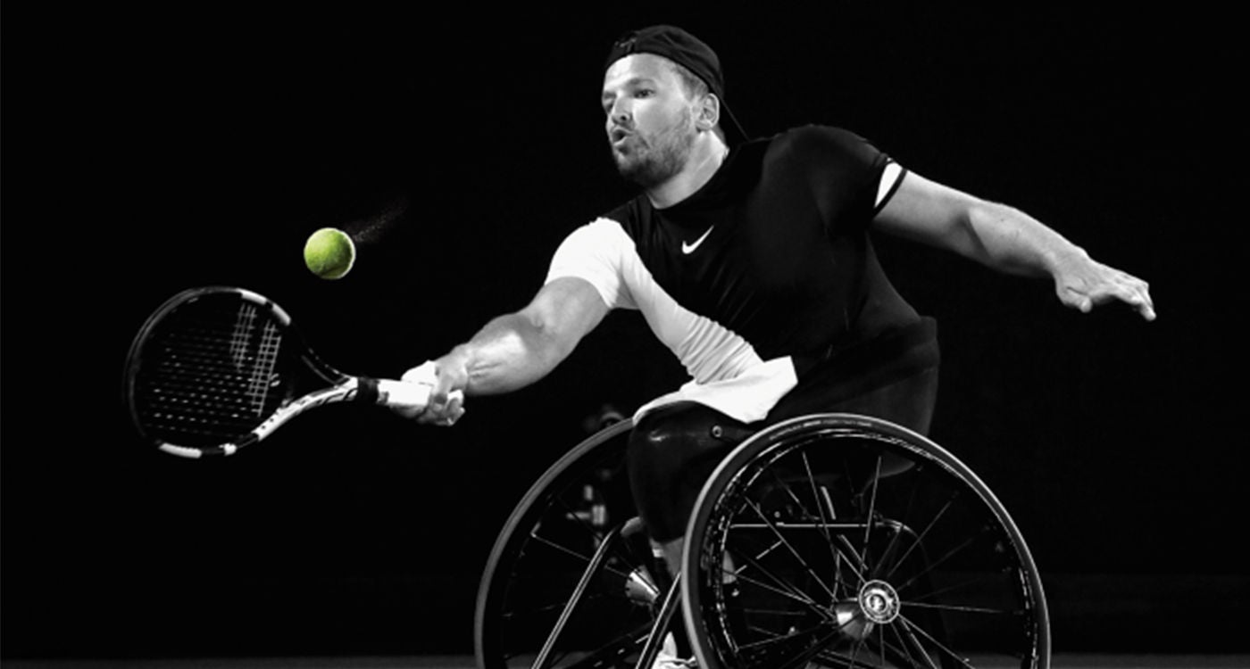 An athlete in a wheelchair is captured mid-swing during a tennis match, focusing on the dynamic movement and intensity of the sport. The player is wearing a two-tone athletic outfit and is positioned to hit a tennis ball with a racket. The scene is set against a dark background, emphasizing the action and the wheelchair's design. The image is in black and white, highlighting contrast and energy.