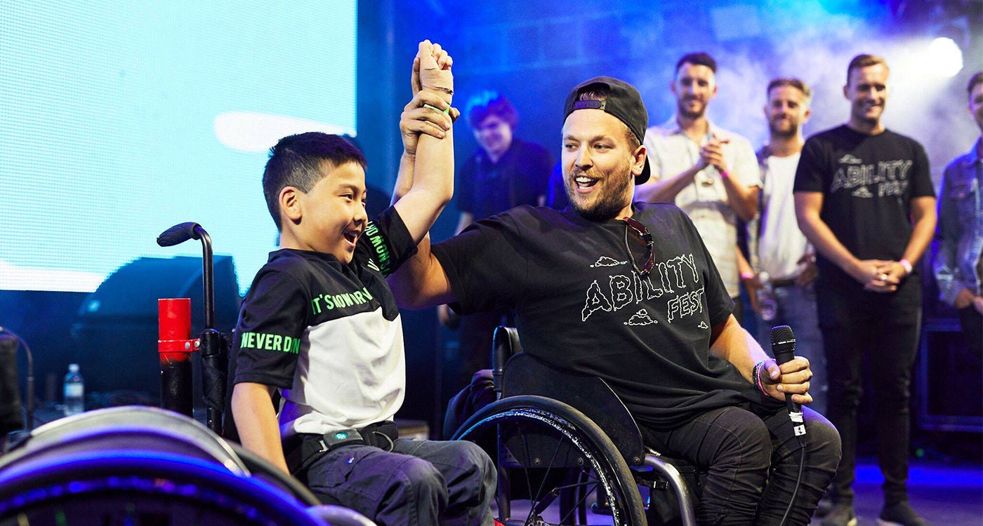 A young boy and an adult, both using wheelchairs, raise their joined hands in a gesture of triumph on a stage. The setting features dramatic lighting and a group of people standing in the background, suggesting a public or motivational event. The adult's shirt displays the word 'ABILITY' in large letters, while the boy's sleeve reads 'NEVER'. The atmosphere is energetic and empowering, with a focus on inclusion and achievement.