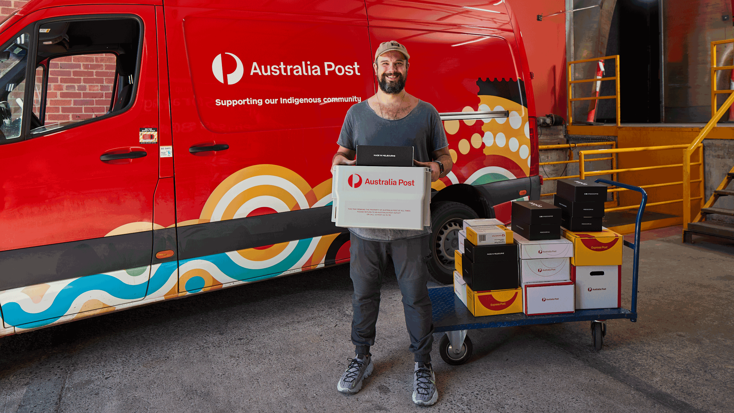 A person stands in front of a red Australia Post van, holding a parcel box. The van features colorful Indigenous artwork and clear Australia Post branding. Multiple packages are stacked on a blue trolley beside the worker. The setting appears to be an industrial loading area with concrete flooring and yellow stairs.