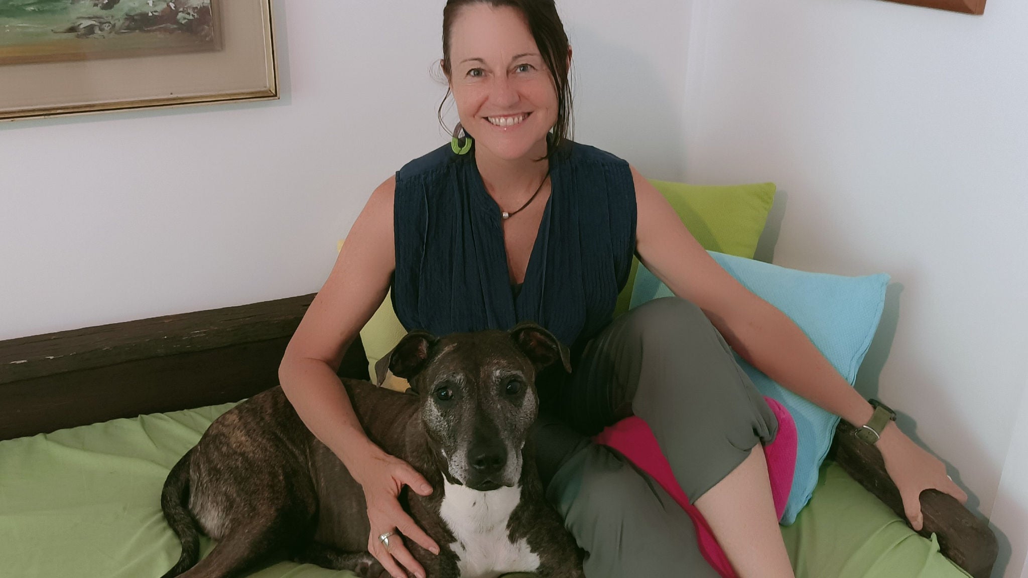 A woman is seated on a bed, gently holding a medium-sized brindle dog. The setting appears to be a cozy, lived-in room with colorful pillows and framed artwork on the wall. The overall mood is relaxed and homey, with soft, natural lighting and casual attire. No visible text or numbers are present in the image.