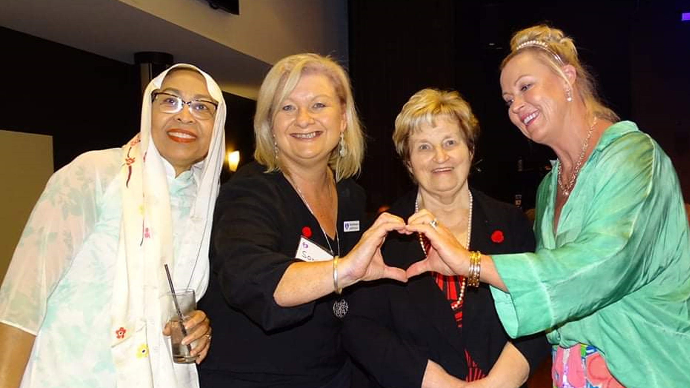Four women are gathered together indoors, with two of them forming a heart shape with their hands. The setting appears to be a social or formal event, as indicated by their attire and name tags. One woman holds a drink, and all are wearing decorative pins. The background is dimly lit, suggesting an evening or indoor venue.