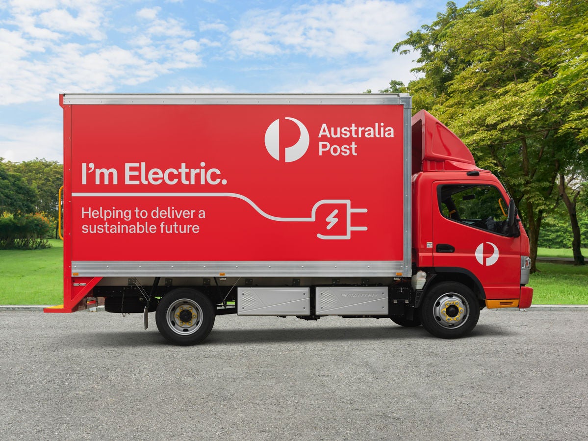 A bright red Australia Post delivery truck is parked on a paved surface in front of a green park setting. Large white text on the side reads "I'm Electric." along with a plug and battery icon, highlighting its electric power. Additional text states "Helping to deliver a sustainable future," emphasizing environmental benefits. Lush trees and grass in the background contrast with the vivid red vehicle, creating a clean and modern look.