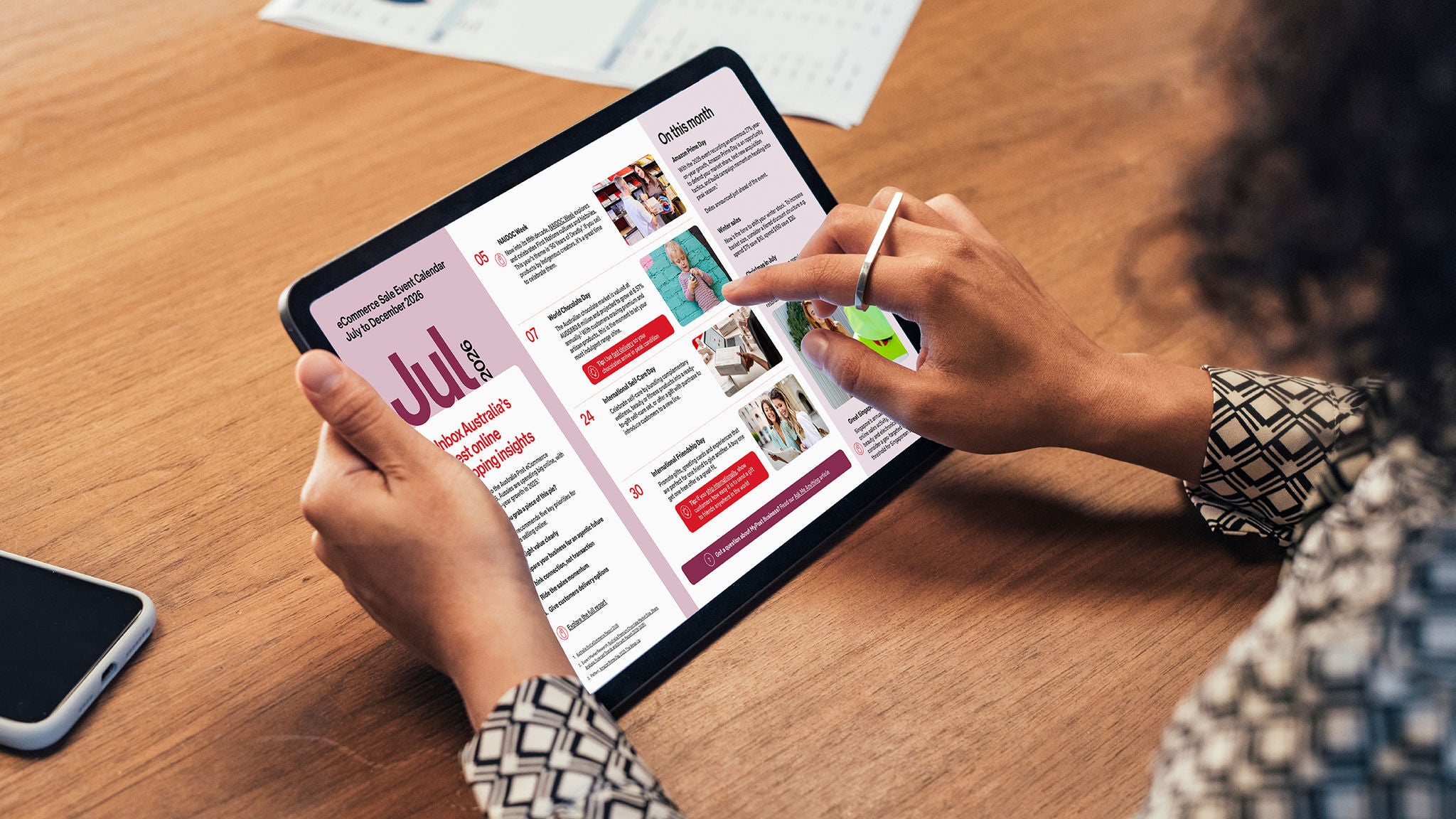 A woman uses a stylus to read a digital newsletter on a tablet at a wooden desk. The tablet screen shows a colorful layout with the month "Jul" and various articles and images. Her other hand holds the device while she scrolls through the content. The scene suggests casual work or personal reading in a modern, tech-savvy environment.