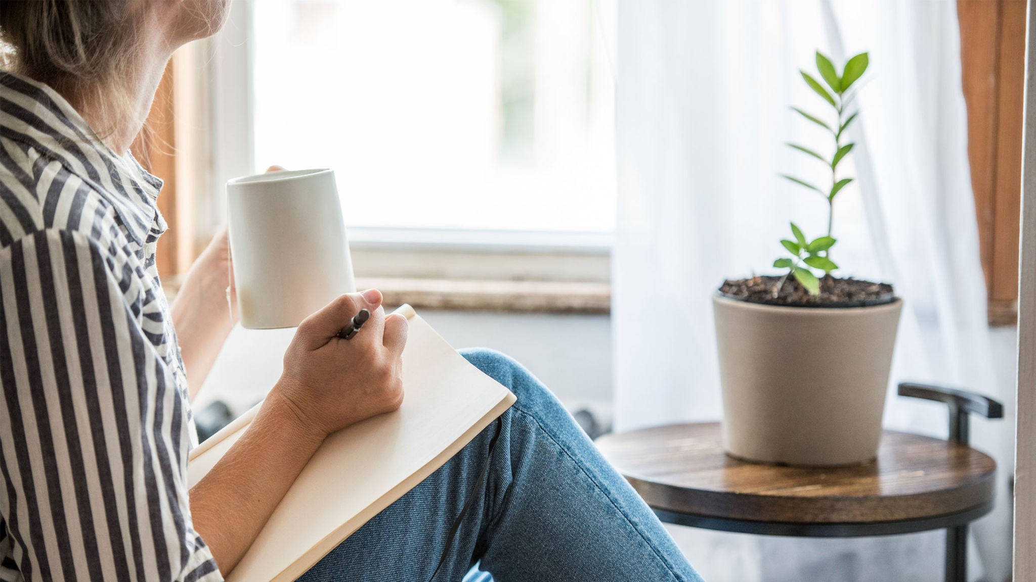 A woman sits indoors near a window, holding a white mug and writing in a notebook. She is dressed casually in a striped shirt and jeans, with a potted plant on a small table beside her. The scene is bright and calm, with natural light filtering through sheer curtains.