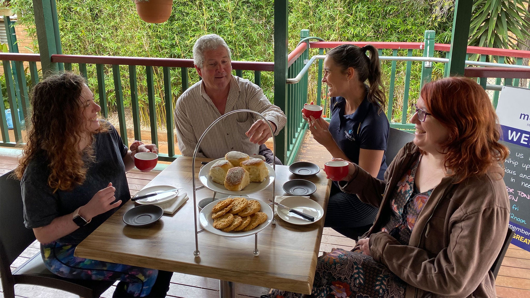 Four adults are seated around a wooden table on a covered outdoor patio, sharing a selection of pastries and hot drinks. The table features a two-tiered tray with scones and cookies, along with red coffee cups and saucers. The setting is casual and relaxed, with greenery visible in the background and a signboard partially visible to the side. The overall mood is warm and social, with natural daylight and earthy tones.