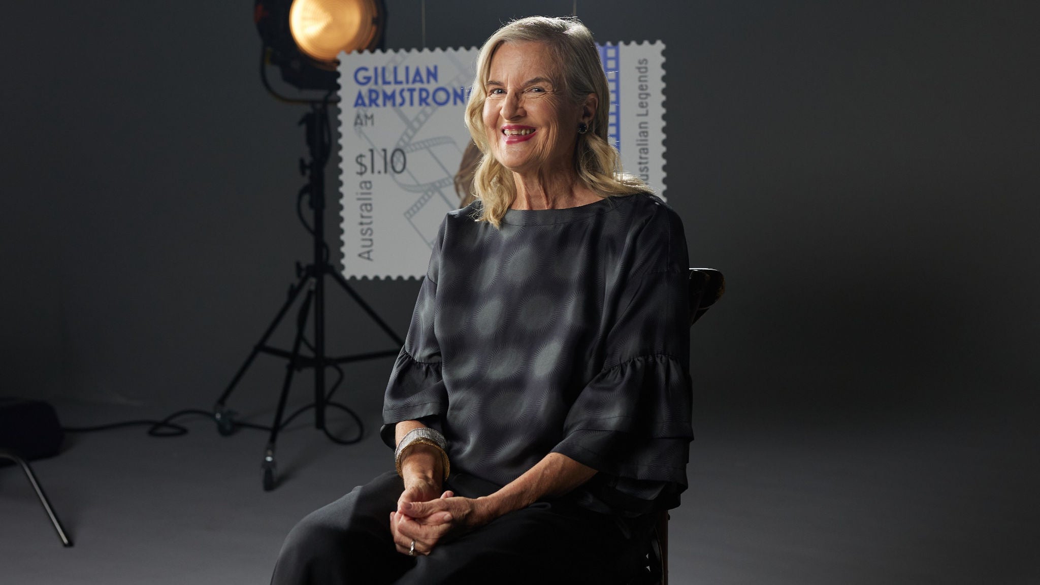 A woman sits on a chair in a dimly lit studio setting. Behind her, a large backdrop features a postage stamp with the text 'Gillian Armstrong', 'Australia', and '$1.10'. Studio lighting equipment is visible, contributing to a professional and creative atmosphere.