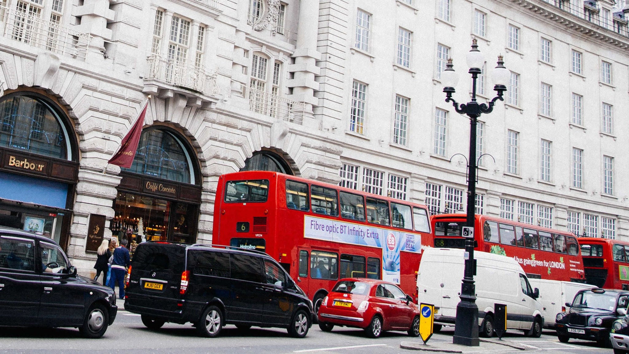 A bustling city street scene features iconic red double-decker buses and various vehicles, including black taxis and white vans, in front of a grand white stone building. Pedestrians walk along the sidewalk near arched shop entrances, with visible signage such as 'Barbo' and 'Optical Express.' The setting is urban, with classic street lamps and a lively atmosphere.
