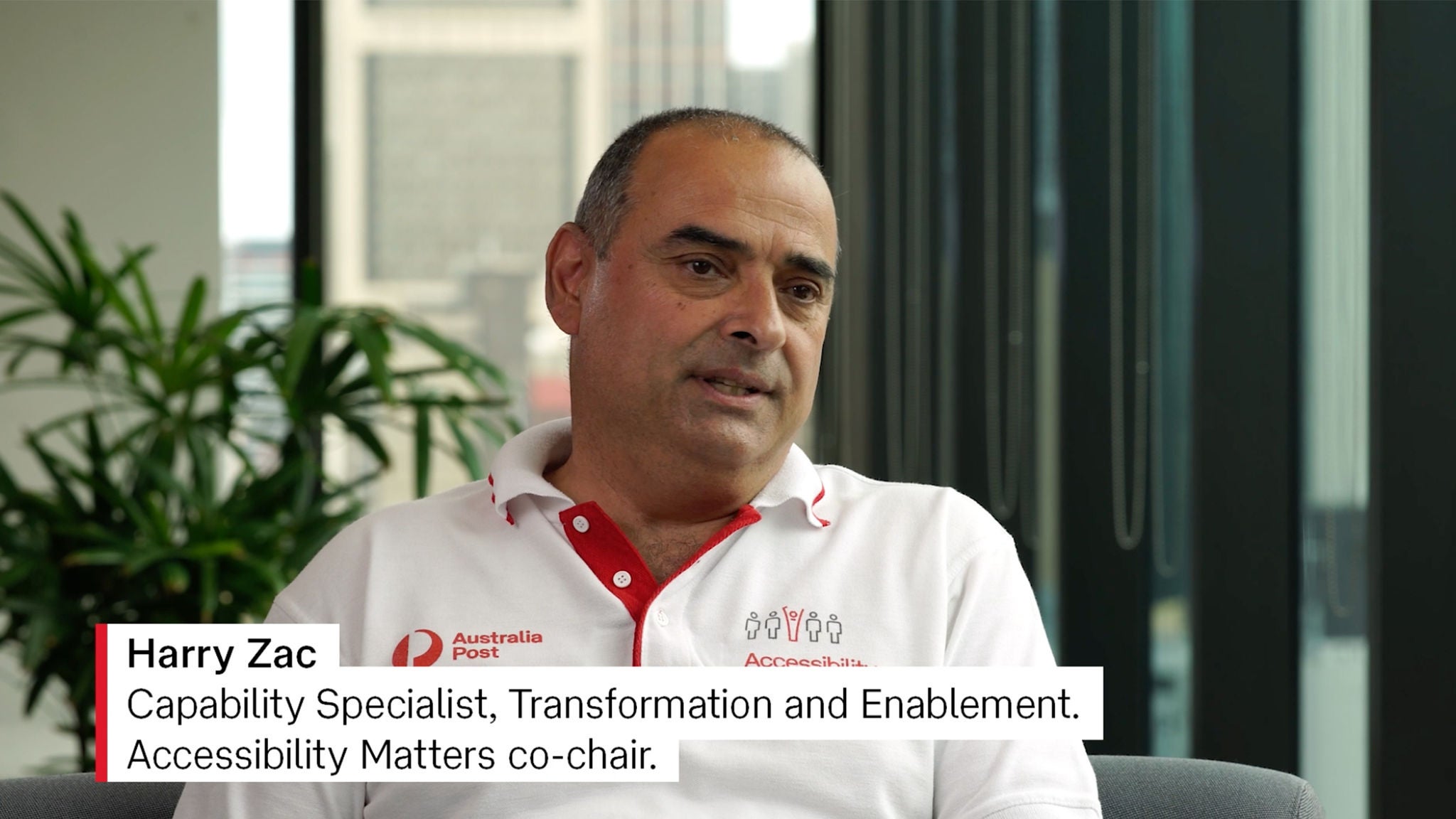 A person wearing a white Australia Post polo shirt is seated indoors for an interview. The setting features large windows and office plants, suggesting a professional environment. The shirt displays the Australia Post logo and accessibility-themed graphics. The text overlay introduces the individual as a Capability Specialist and Accessibility Matters co-chair.