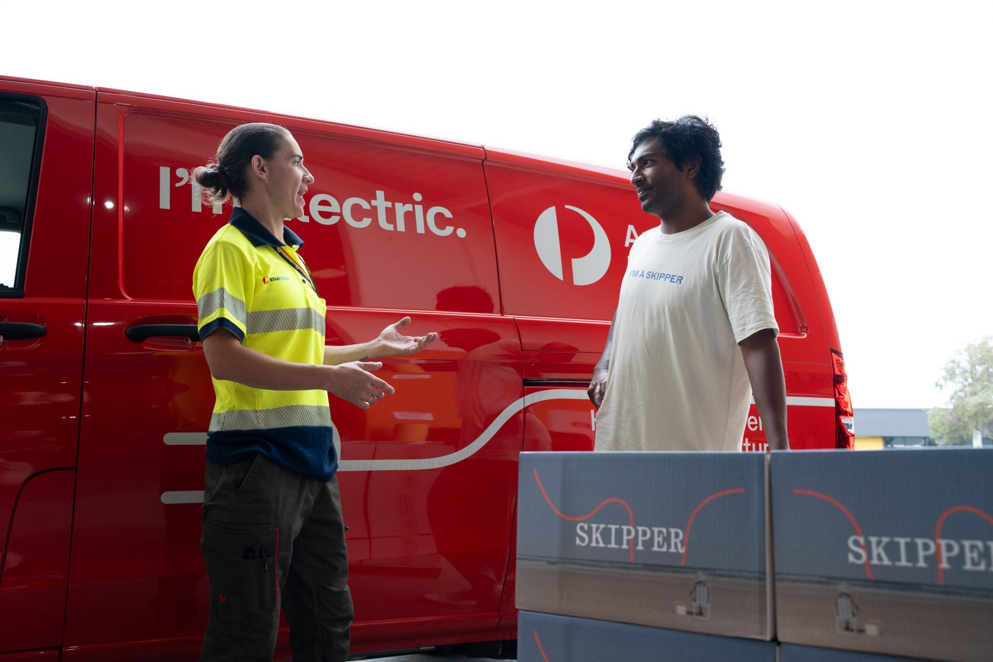 A courier in a high-visibility uniform talks with a customer beside a bright red electric delivery van. Several large boxes labeled "SKIPPER" are stacked in the foreground, suggesting an active parcel drop-off or pickup. The scene appears to take place at a loading area or warehouse entrance during daytime. The overall mood is professional and focused on modern, eco-friendly delivery services.