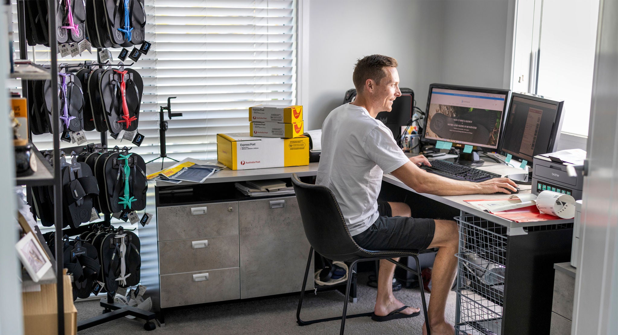 A man sits at a desk in a home office, working on a computer with multiple monitors. The room features organized racks of flip-flops and several yellow and white shipping boxes, suggesting an e-commerce or small business setting. The environment is bright with natural light from a window, and the overall mood is productive and casual. No visible numeric values are present in the image.
