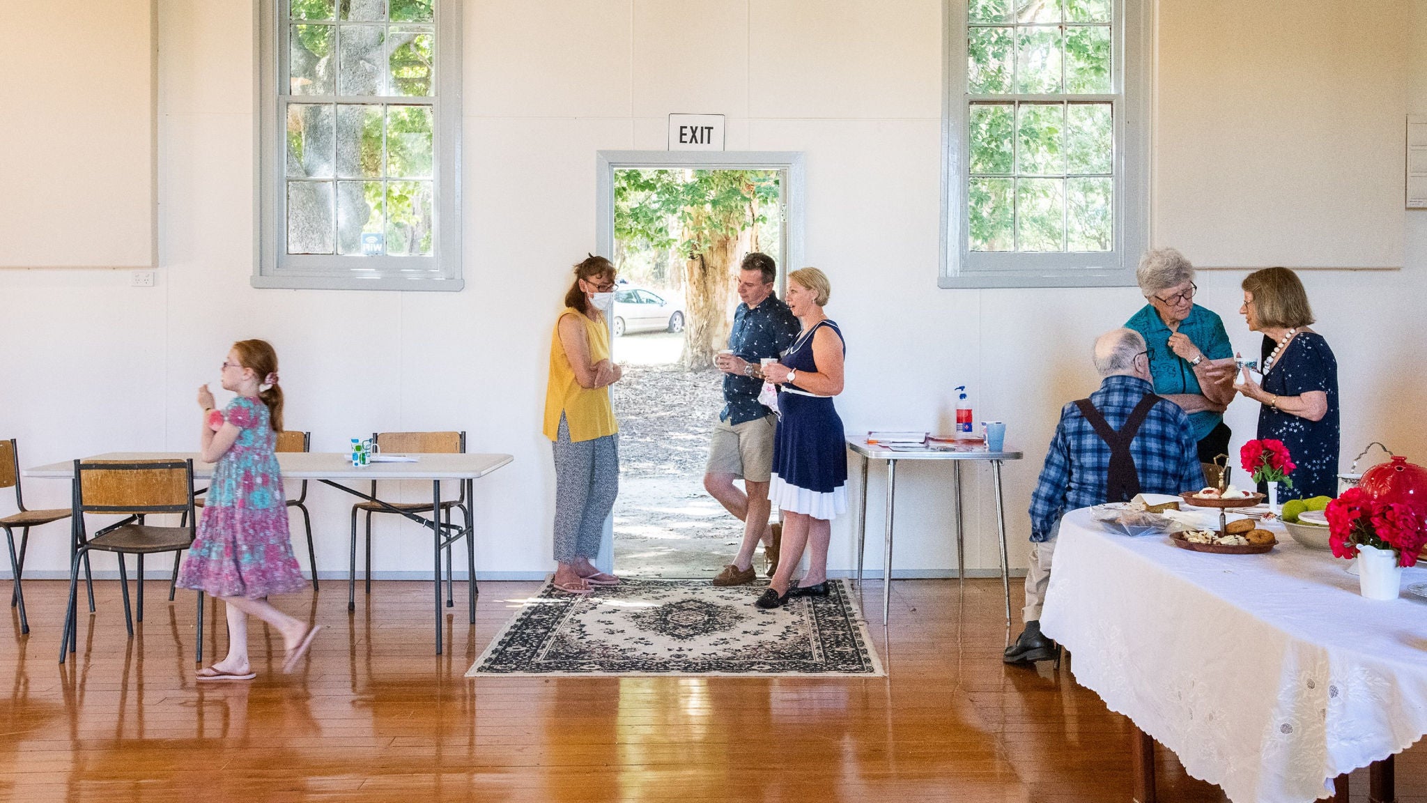 A group of people of various ages socialize in a sunlit community hall. The scene features adults and a child, some standing and chatting while others are seated or serving food from a table with a white tablecloth and red flowers. Large windows let in natural light, and a patterned rug is placed near the exit door. The atmosphere is casual and welcoming, with a mix of light colors and wooden flooring.