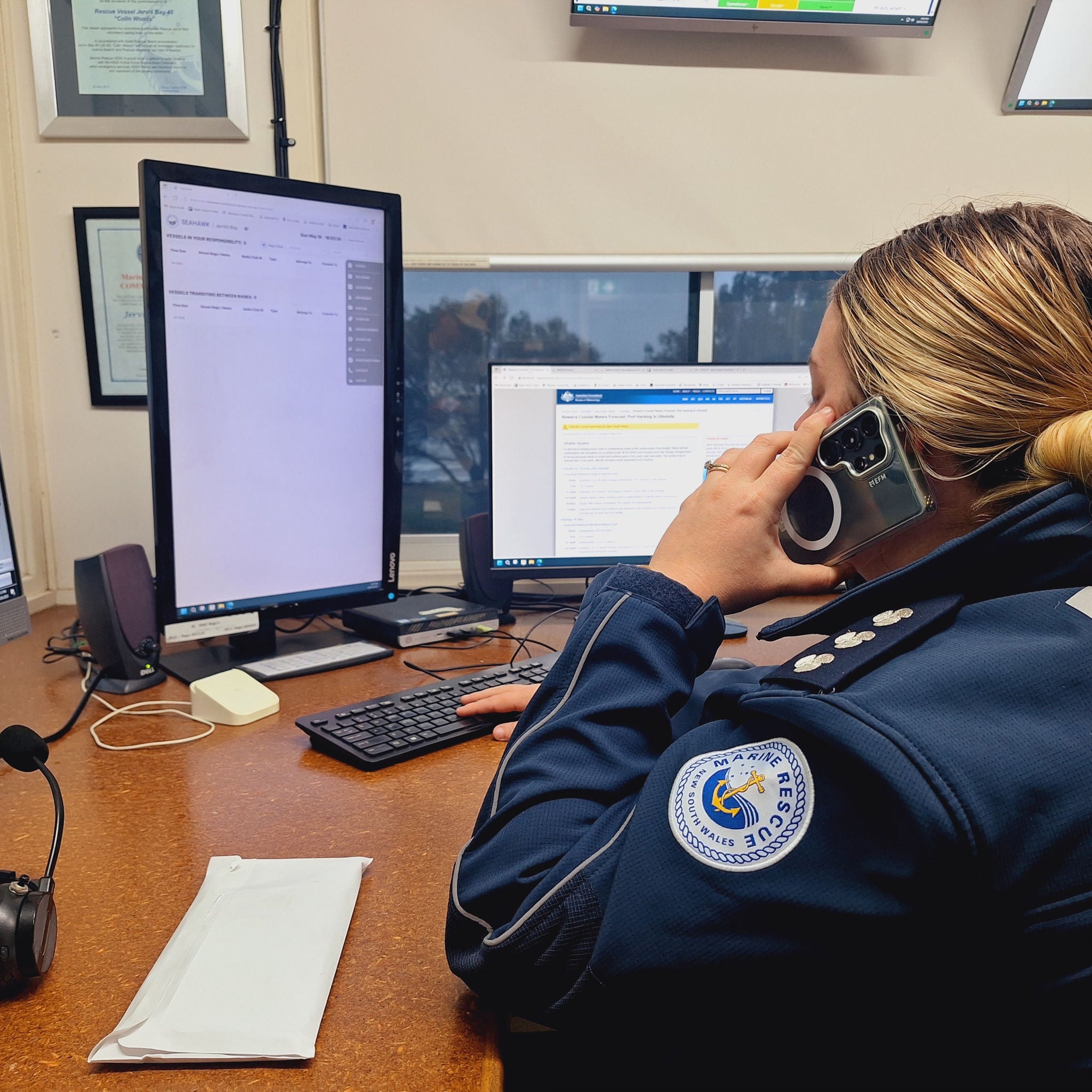 A uniformed emergency dispatcher sits at a desk with multiple computer monitors, speaking on a phone. The setting appears to be a control or dispatch center, with visible screens displaying text and official documents. The person is wearing a navy blue uniform with a visible patch, and the environment is professional and organized.