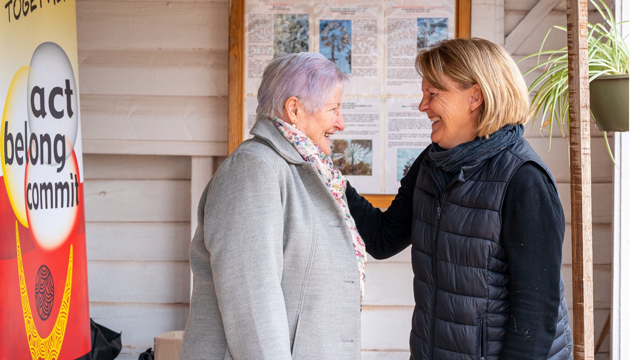 Two adult women are engaged in conversation in front of a wooden wall with a community bulletin board. One woman wears a light gray coat and the other a dark quilted vest. A colorful sign with the words 'act belong commit' is visible, suggesting a community or wellness initiative. The setting appears informal and supportive, with natural light and a potted plant adding warmth.