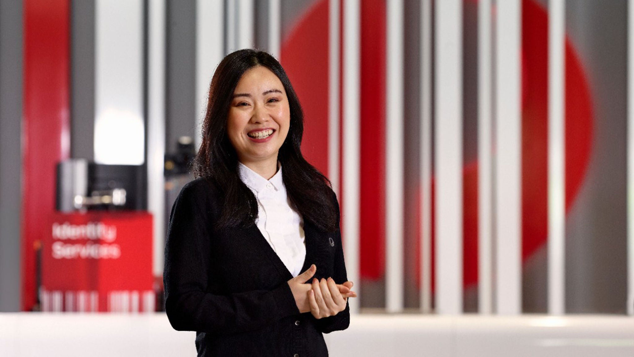 A woman stands in a contemporary office environment featuring vertical striped panels and a large red circle in the background. She is dressed in business attire, including a white shirt and dark cardigan. The setting includes a red sign with the visible text 'Identity Services.' The overall palette is dominated by red, white, and gray tones, creating a professional and modern atmosphere.