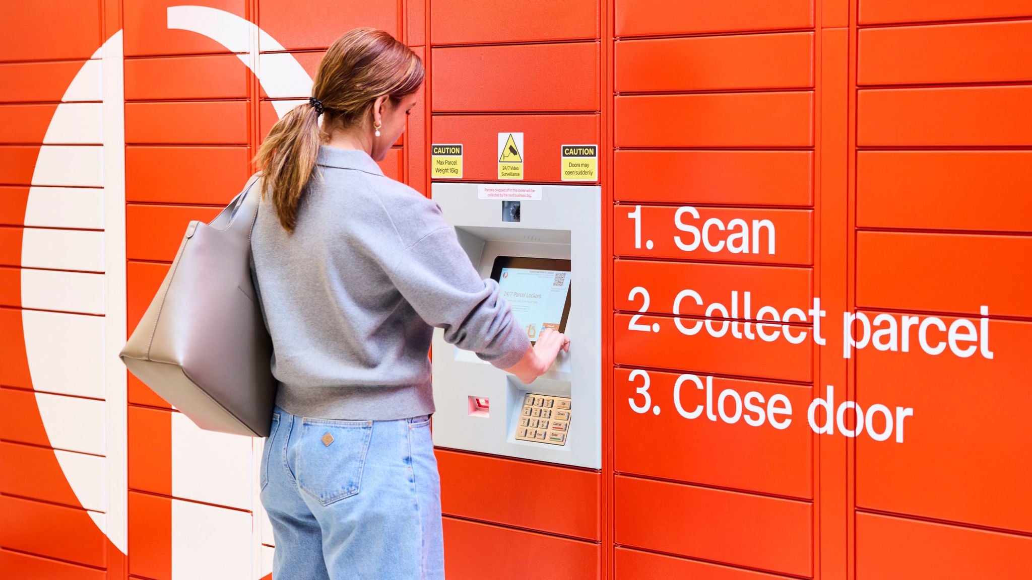 A young woman stands at an orange self-service parcel locker, interacting with the central touchscreen terminal. She holds a large envelope or flat parcel under one arm while operating the machine. Large white instructional text on the lockers reads "1. Scan", "2. Collect parcel", and "3. Close door". The scene suggests a modern, convenient package pickup experience in a brightly colored, indoor or sheltered location.